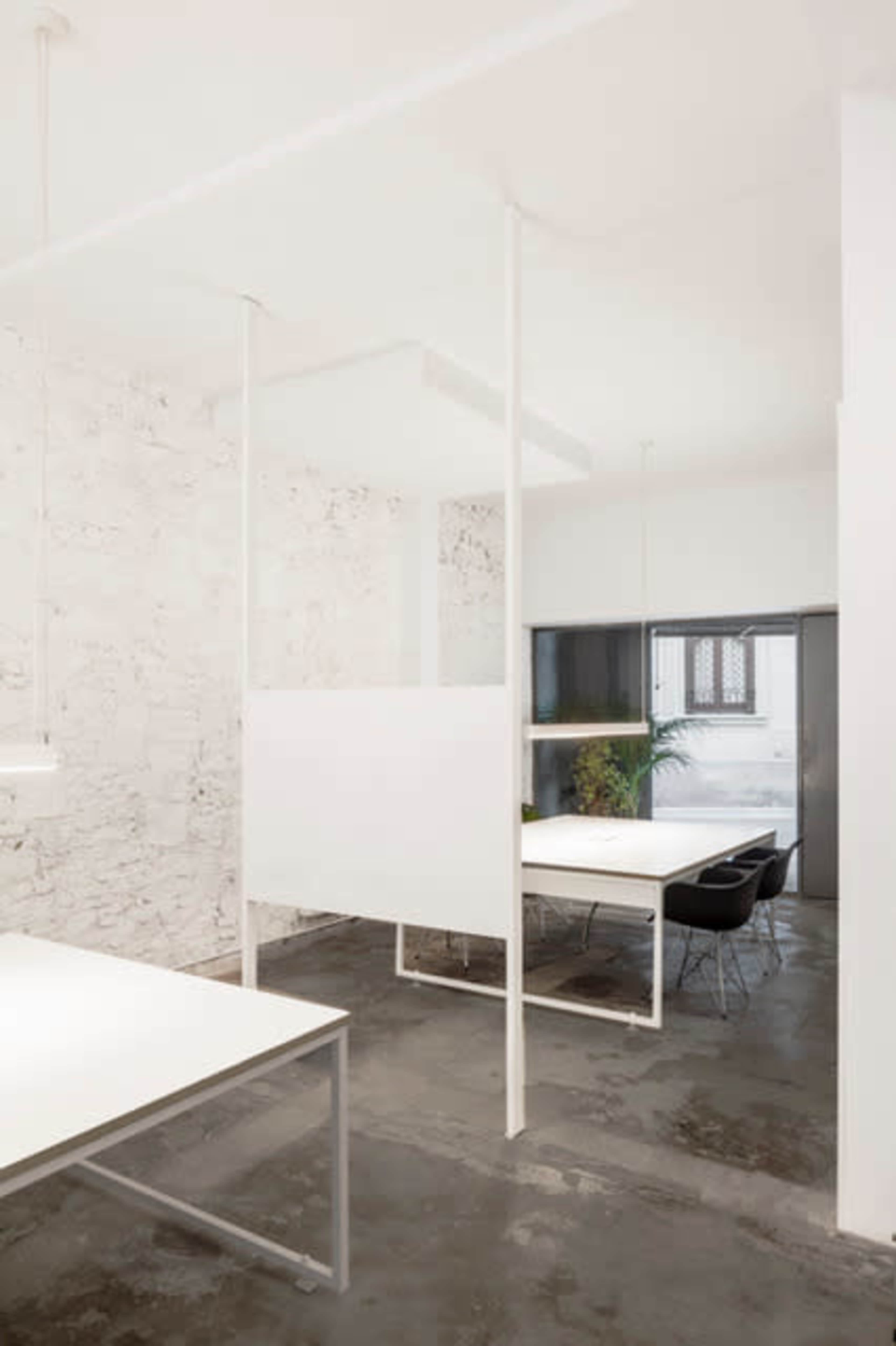 A minimalistic office space featuring white walls, several white desks, and a large window overlooking a green plant.