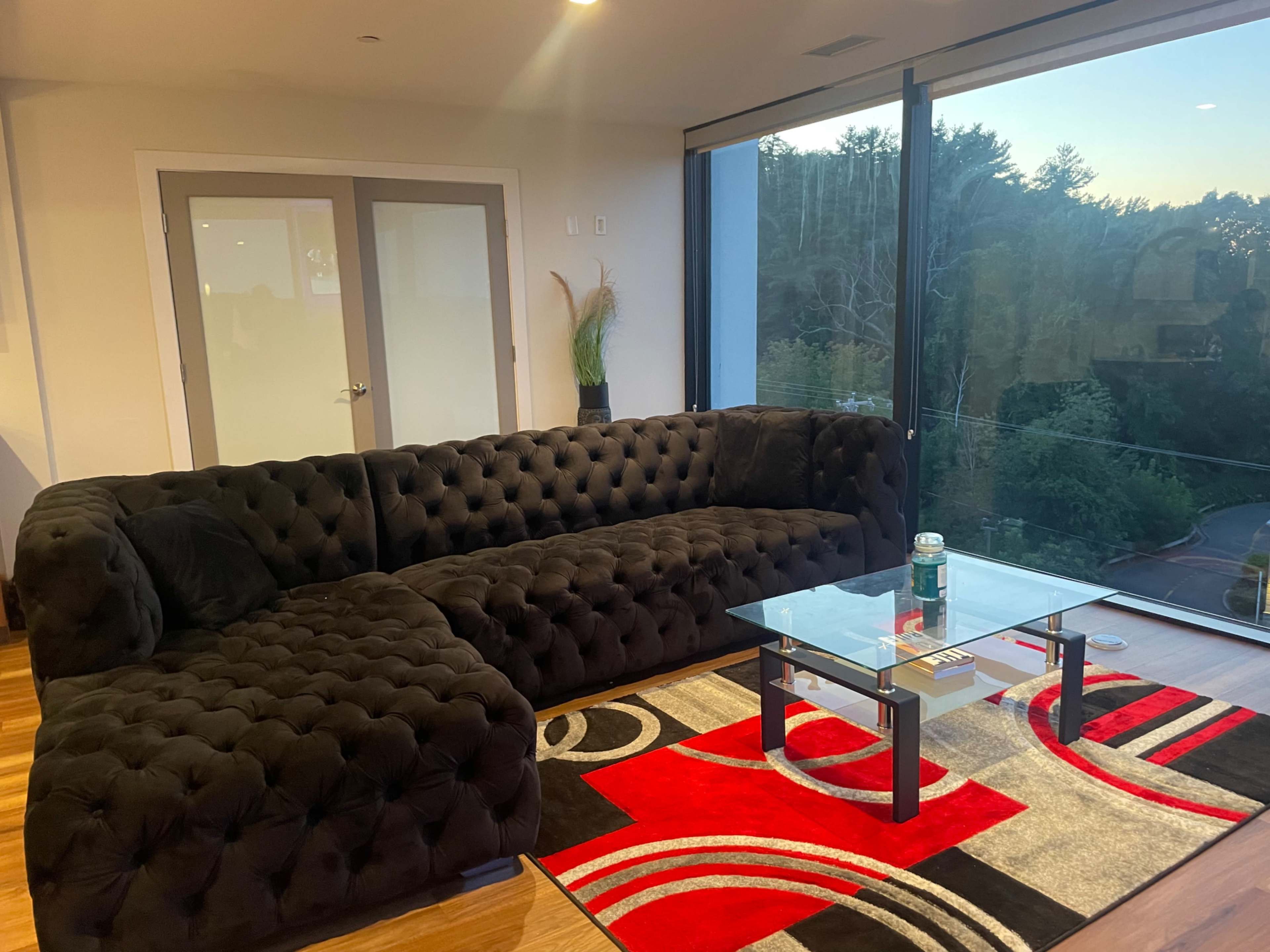 The scene features a black tufted sectional sofa positioned next to a glass coffee table on a patterned rug, with large windows revealing a view of greenery outside.