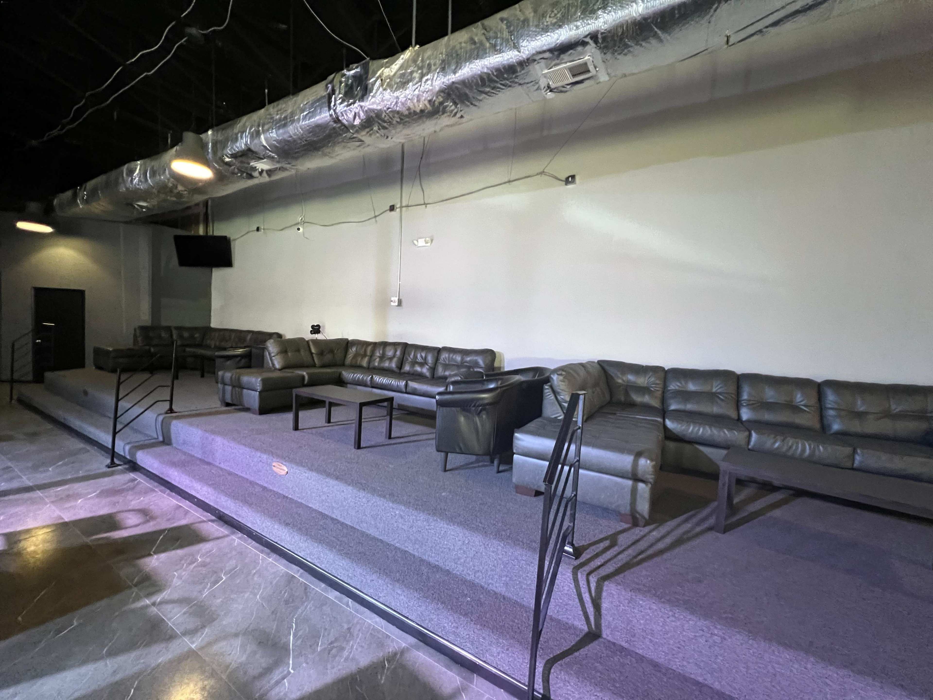 A spacious lounge area featuring multiple leather sofas arranged on a raised platform with a carpeted surface and industrial-style lighting overhead.