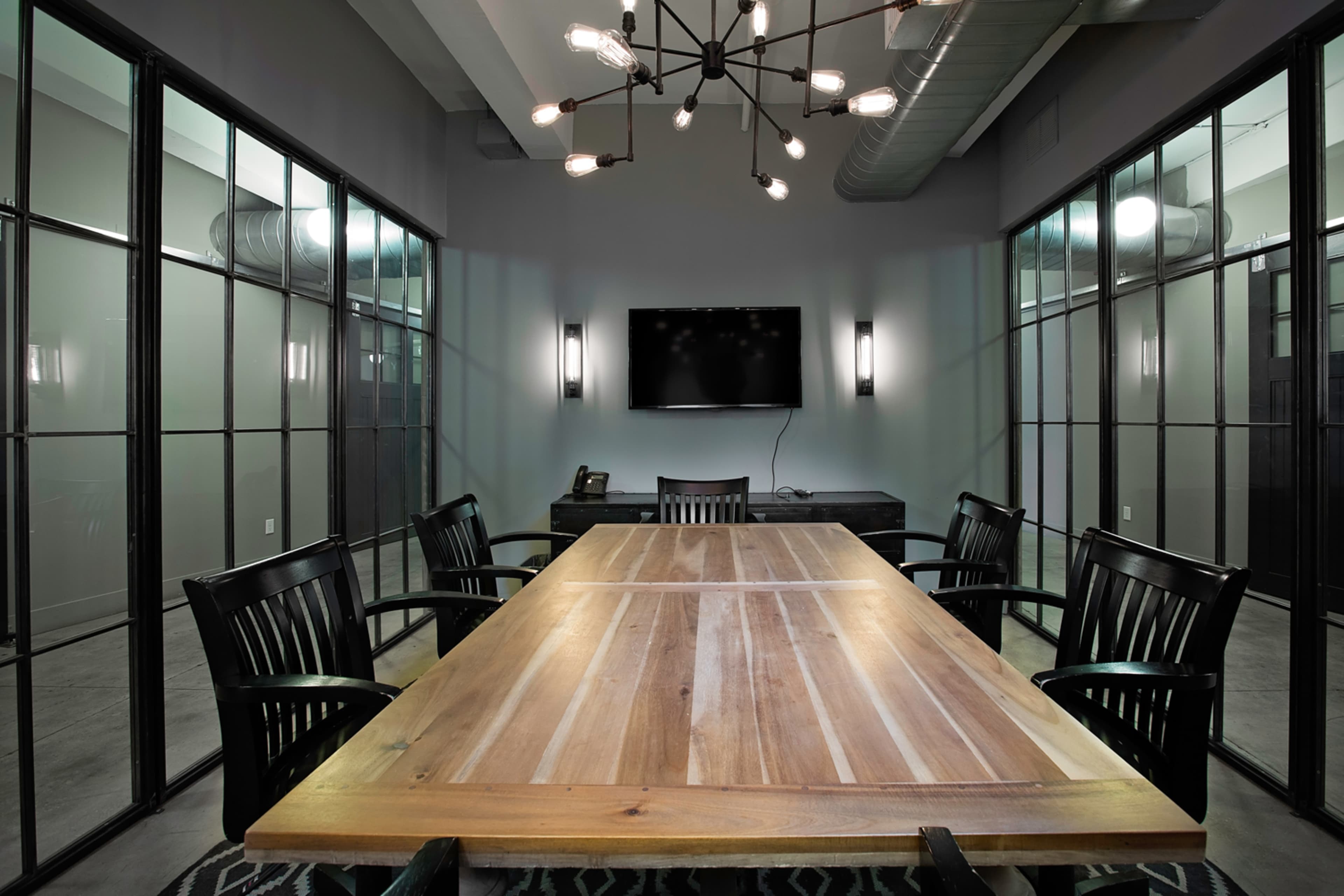A long wooden conference table surrounded by black chairs is situated in a glass-walled meeting room with a large wall-mounted TV.