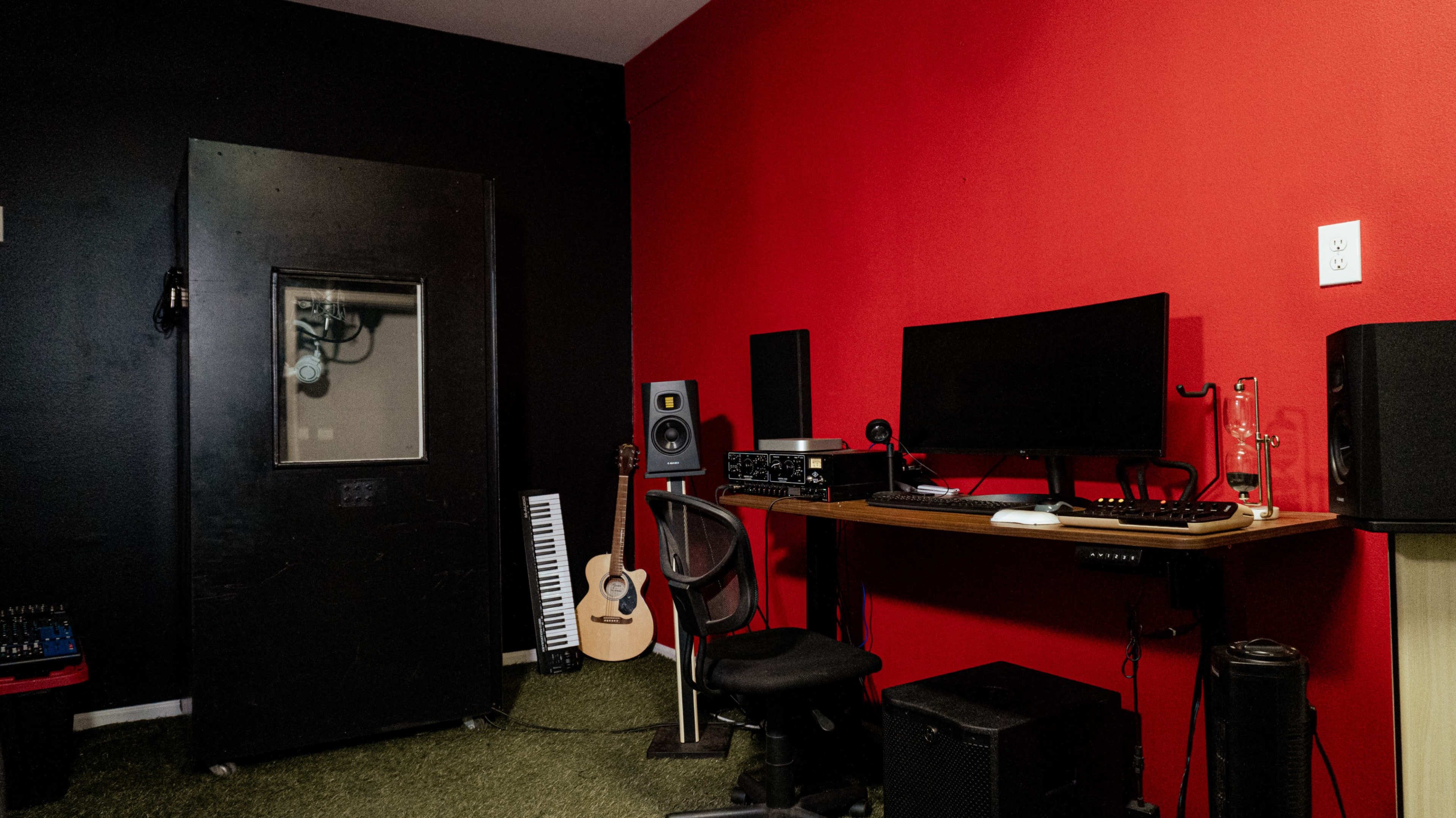 The image shows a music studio corner with a black wall, a red wall, a desk with a computer monitor, audio equipment, an acoustic guitar, and a large door.