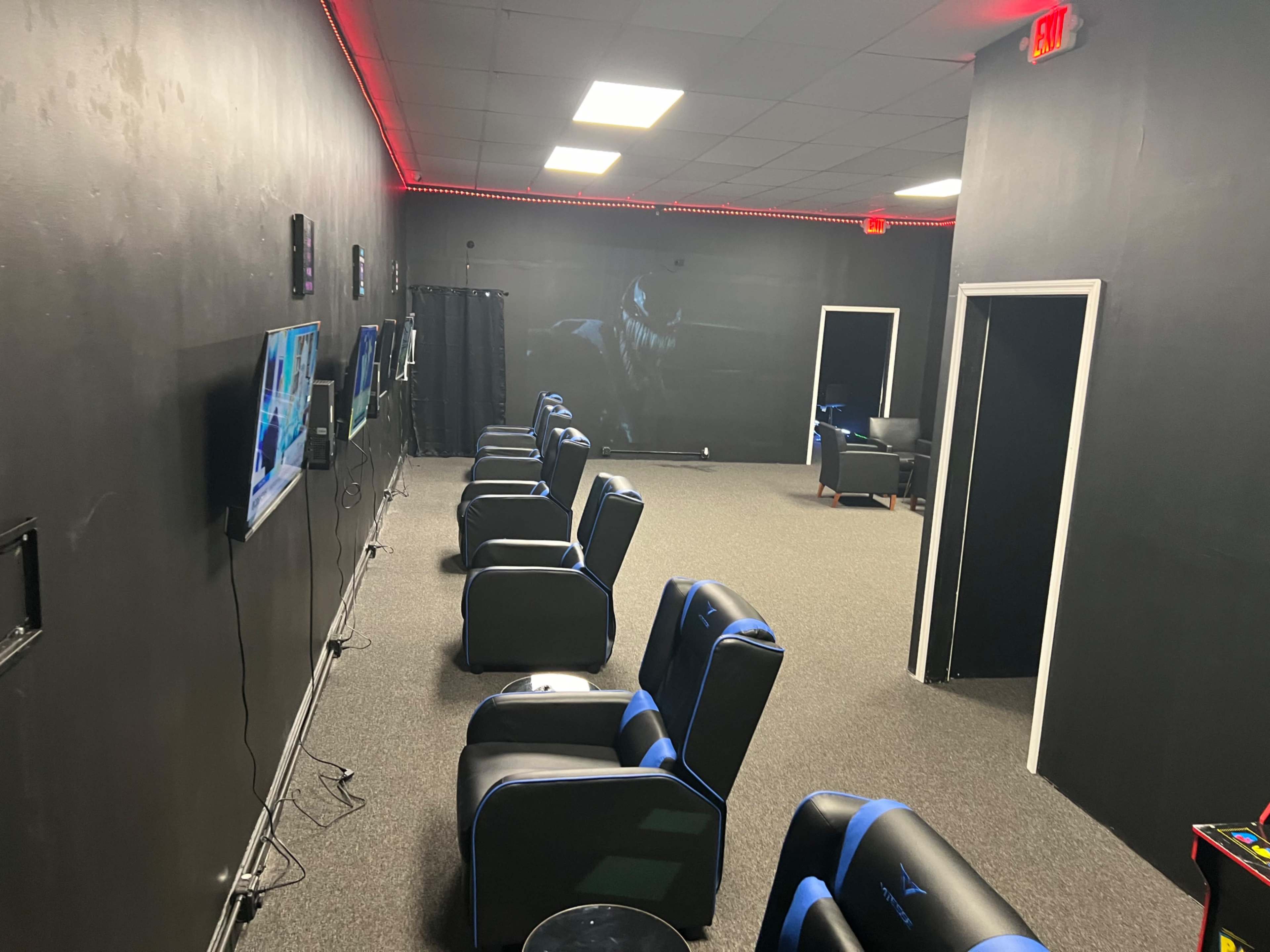 The room features several gaming chairs facing multiple wall-mounted televisions against a black backdrop.