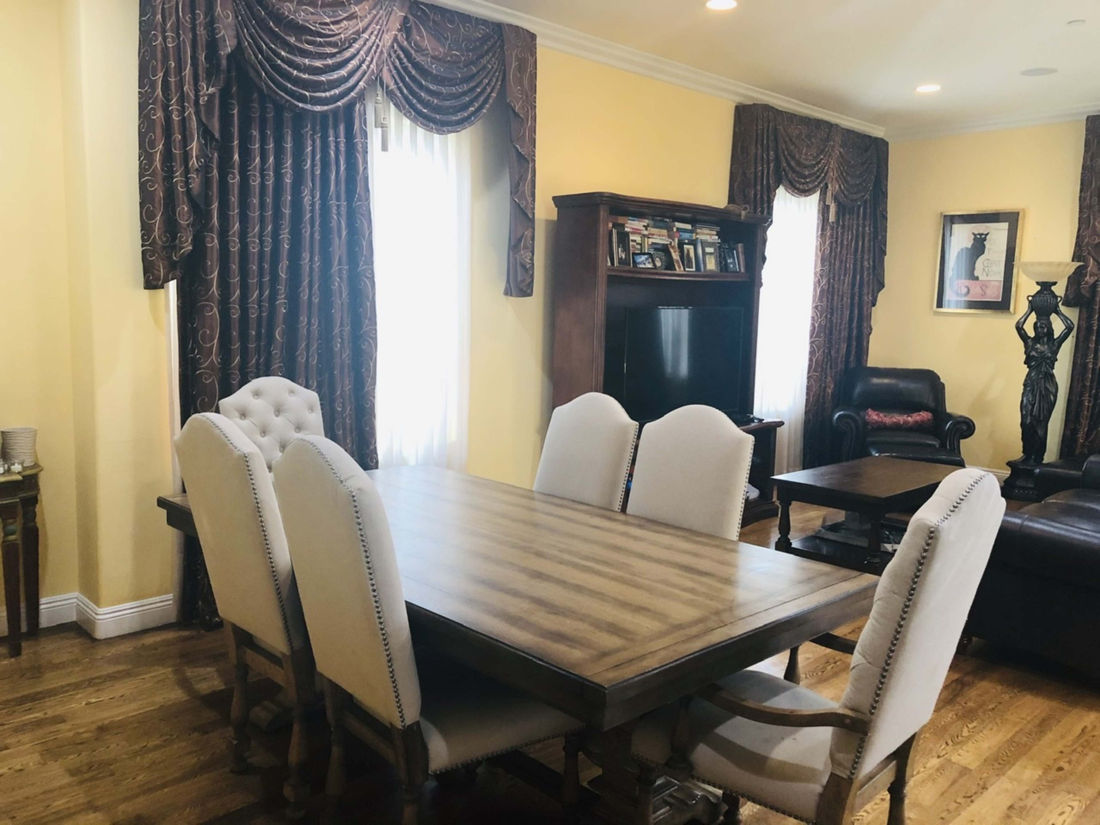 A wooden dining table with six upholstered chairs is positioned in a brightly lit room featuring yellow walls and decorative curtains.