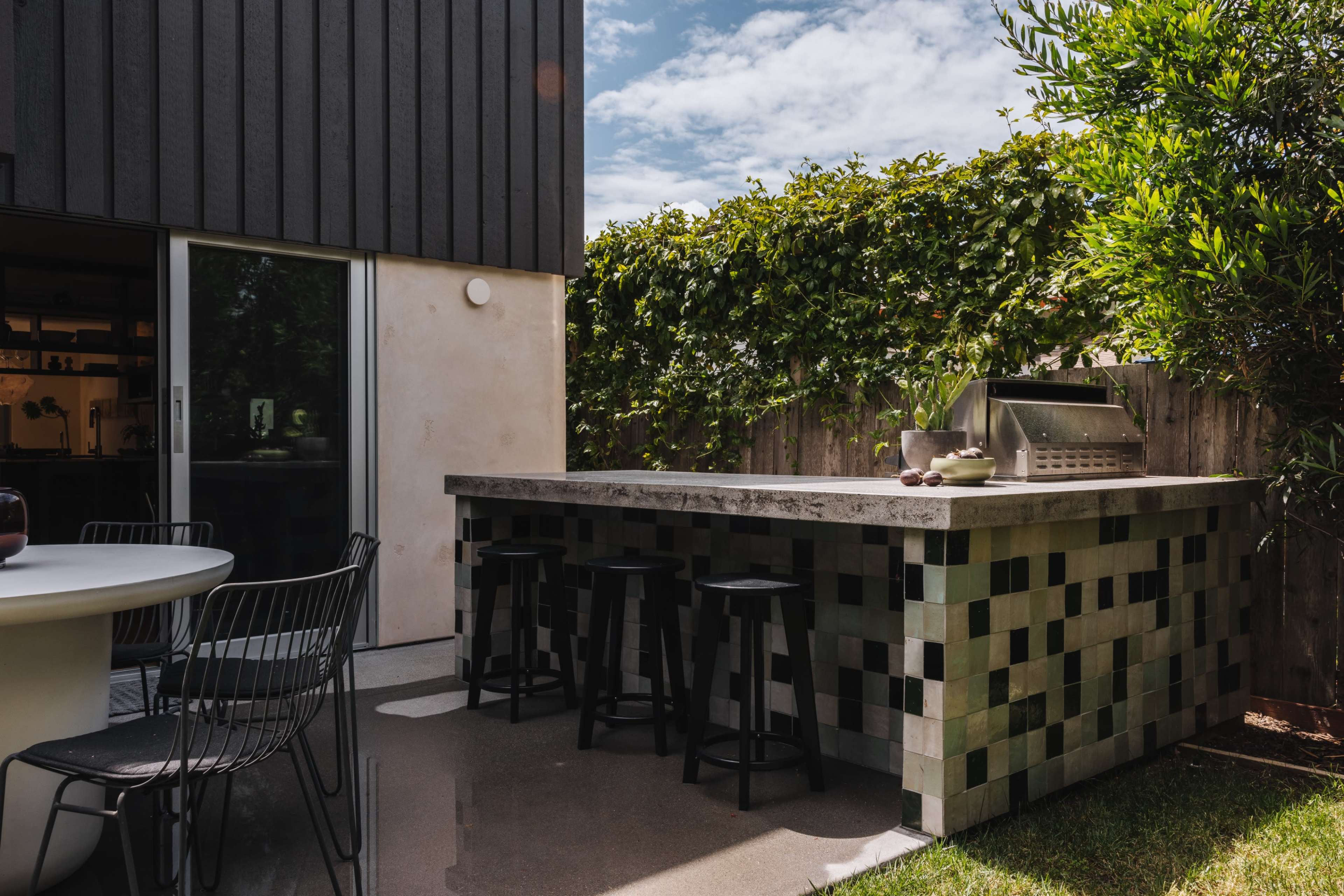 The image shows an outdoor kitchen with a stone countertop and a grill, surrounded by greenery and a modern black building.