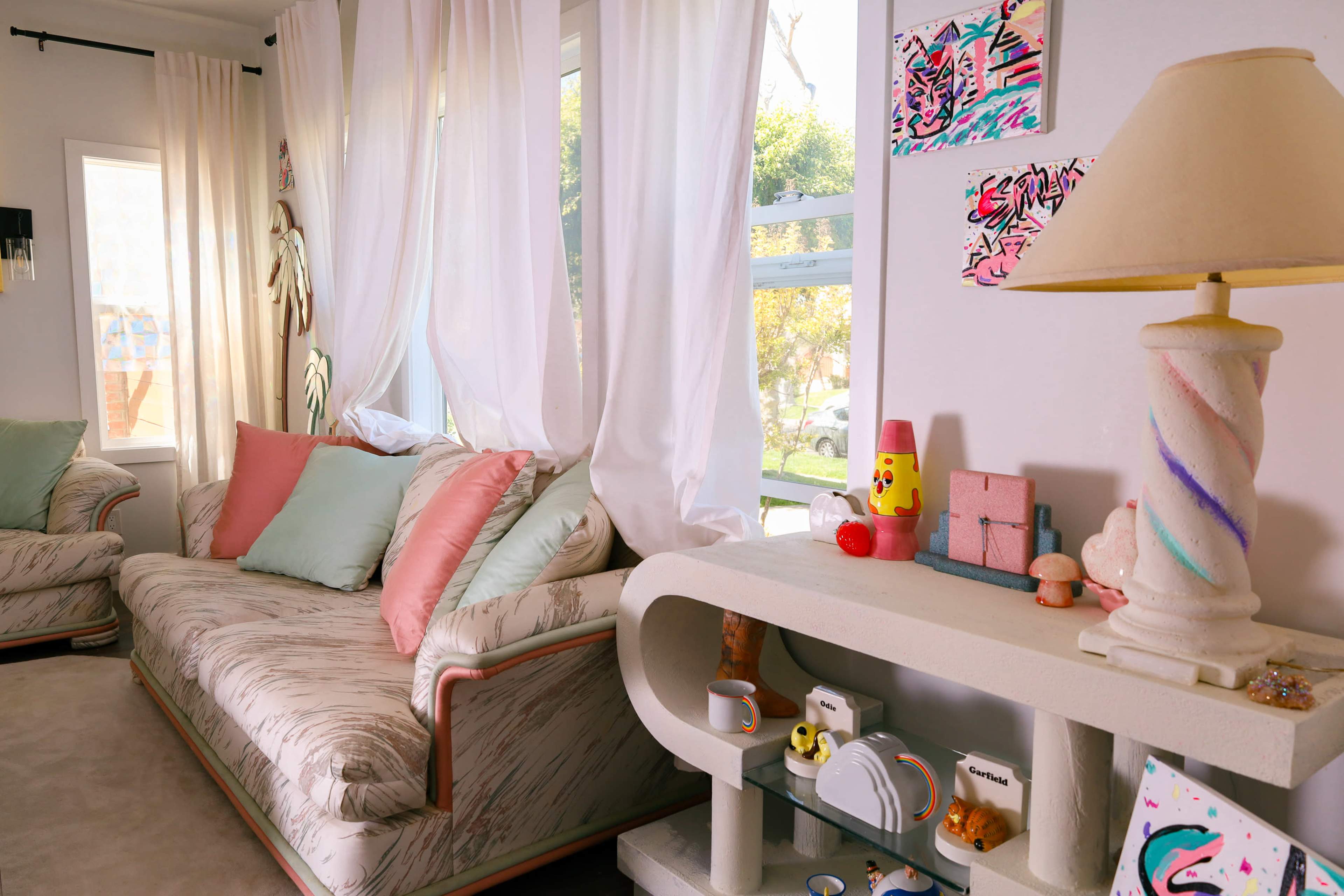 The image shows a cozy living room with a light-colored sofa adorned with pastel cushions, a decorative lamp, and various knick-knacks on a unique console table beside large windows.