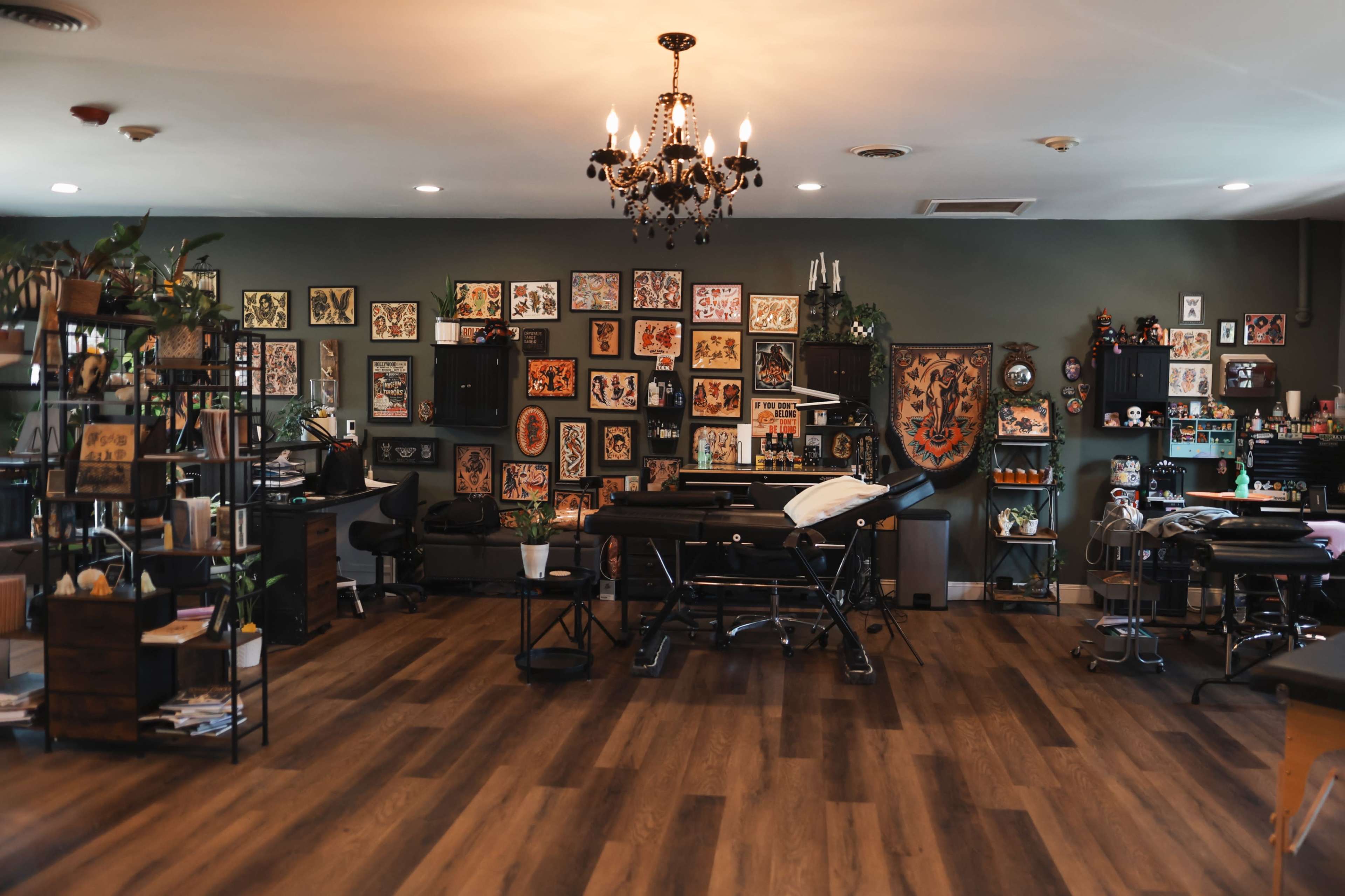 The image shows a tattoo studio with tattoo stations, artwork on the walls, and decorative plants.
