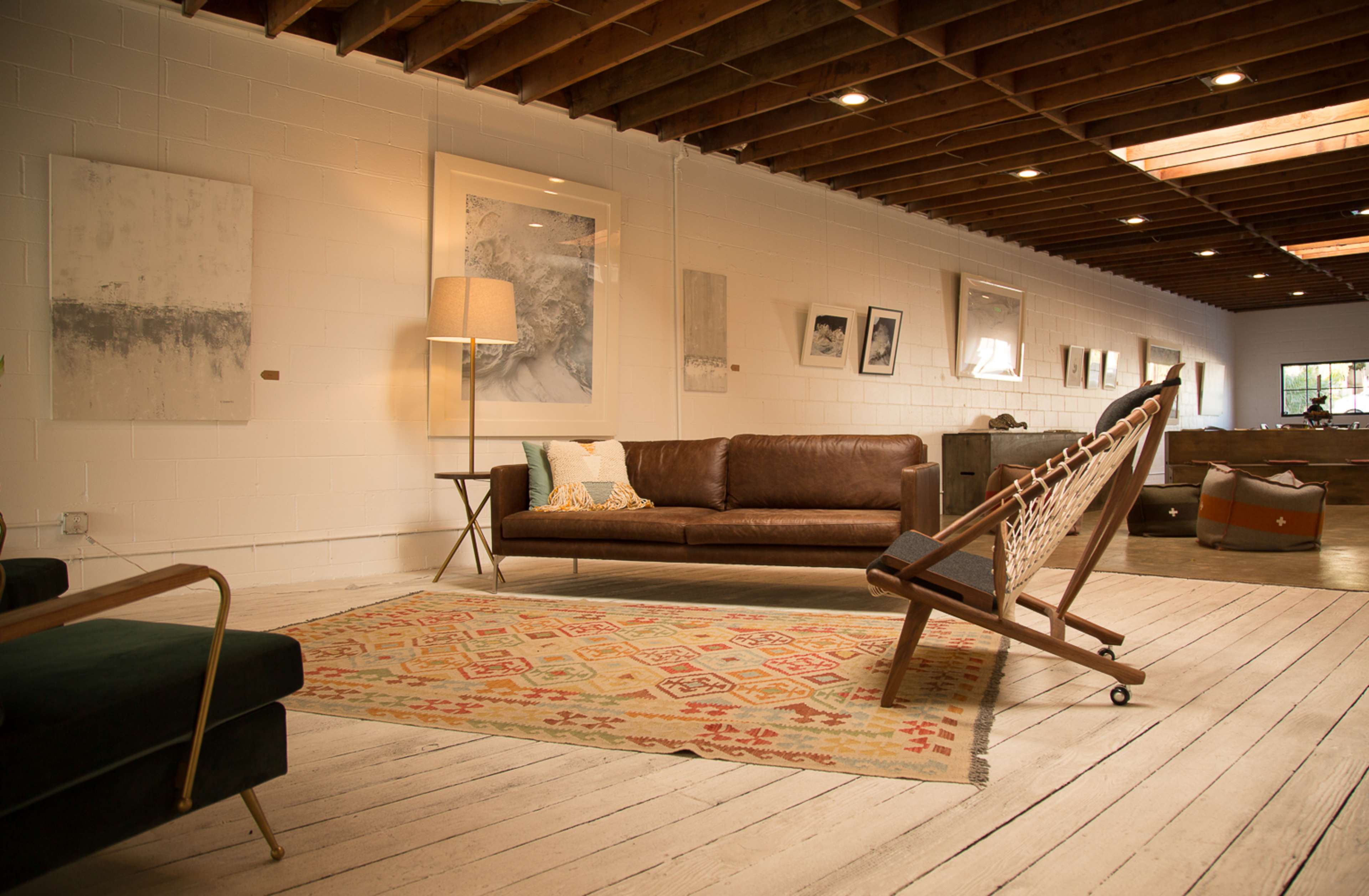 The image shows a spacious living area with a leather sofa, a mid-century lamp, and a wooden rocking chair on a patterned rug, surrounded by various framed artworks on the walls.