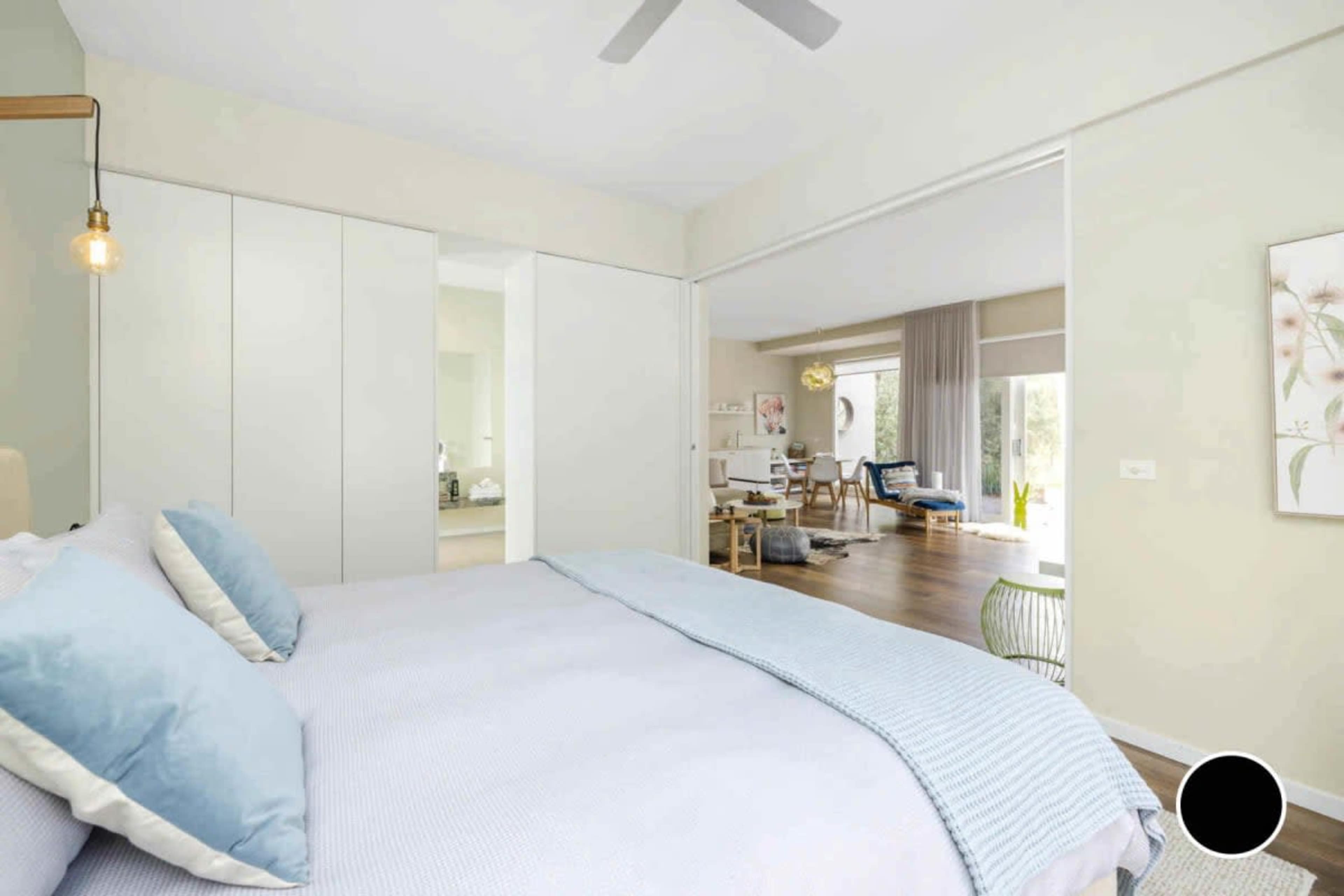 A neatly arranged bedroom features a bed with light blue bedding and an open doorway leading to a sunlit living area.