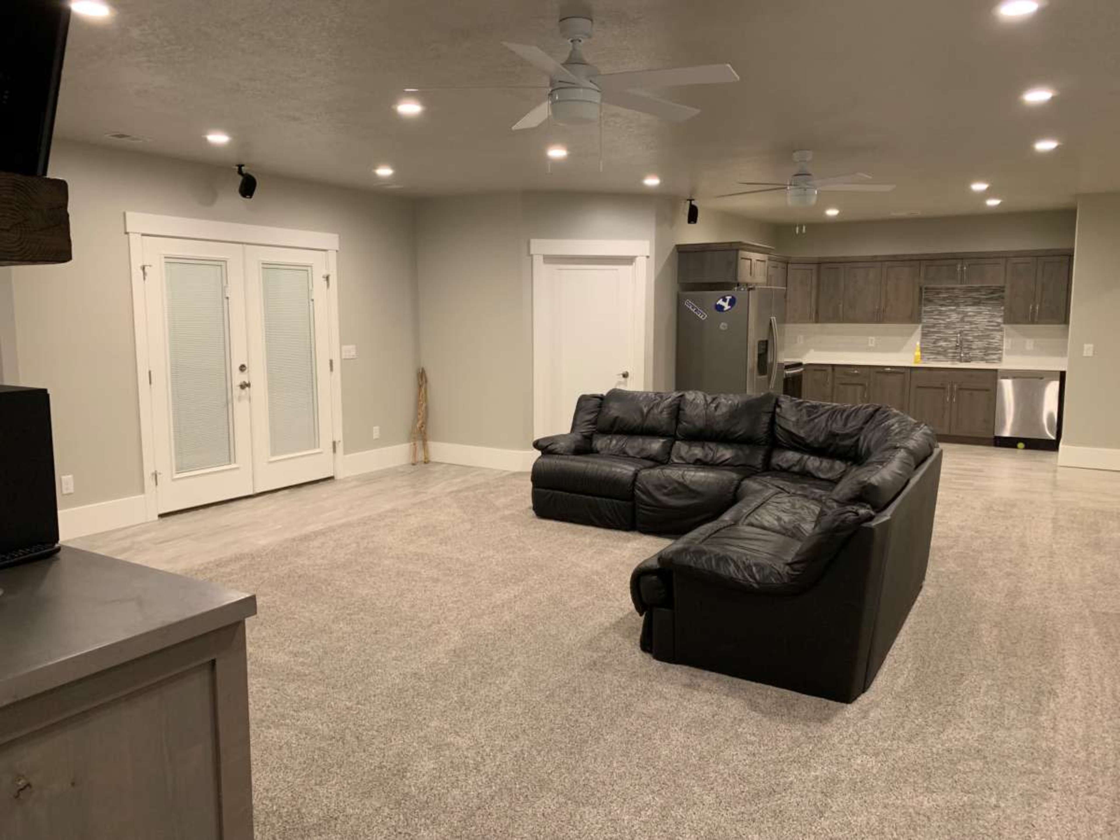 The image shows a spacious living area with a large black sectional sofa, gray carpet, and a modern kitchen in the background.