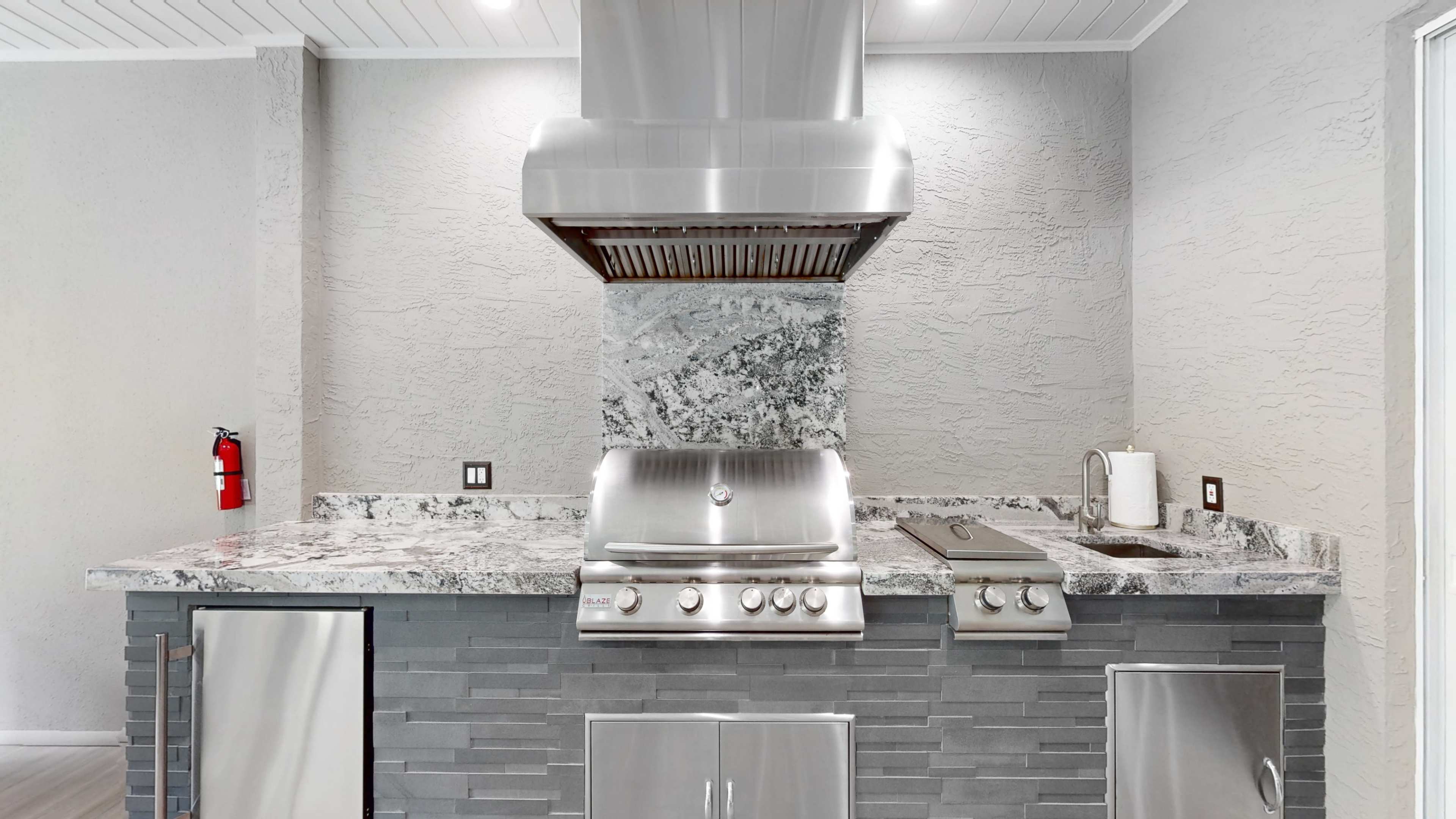 The image shows a modern outdoor kitchen with a stainless steel grill and hood, surrounded by a marble countertop and gray stone walls.