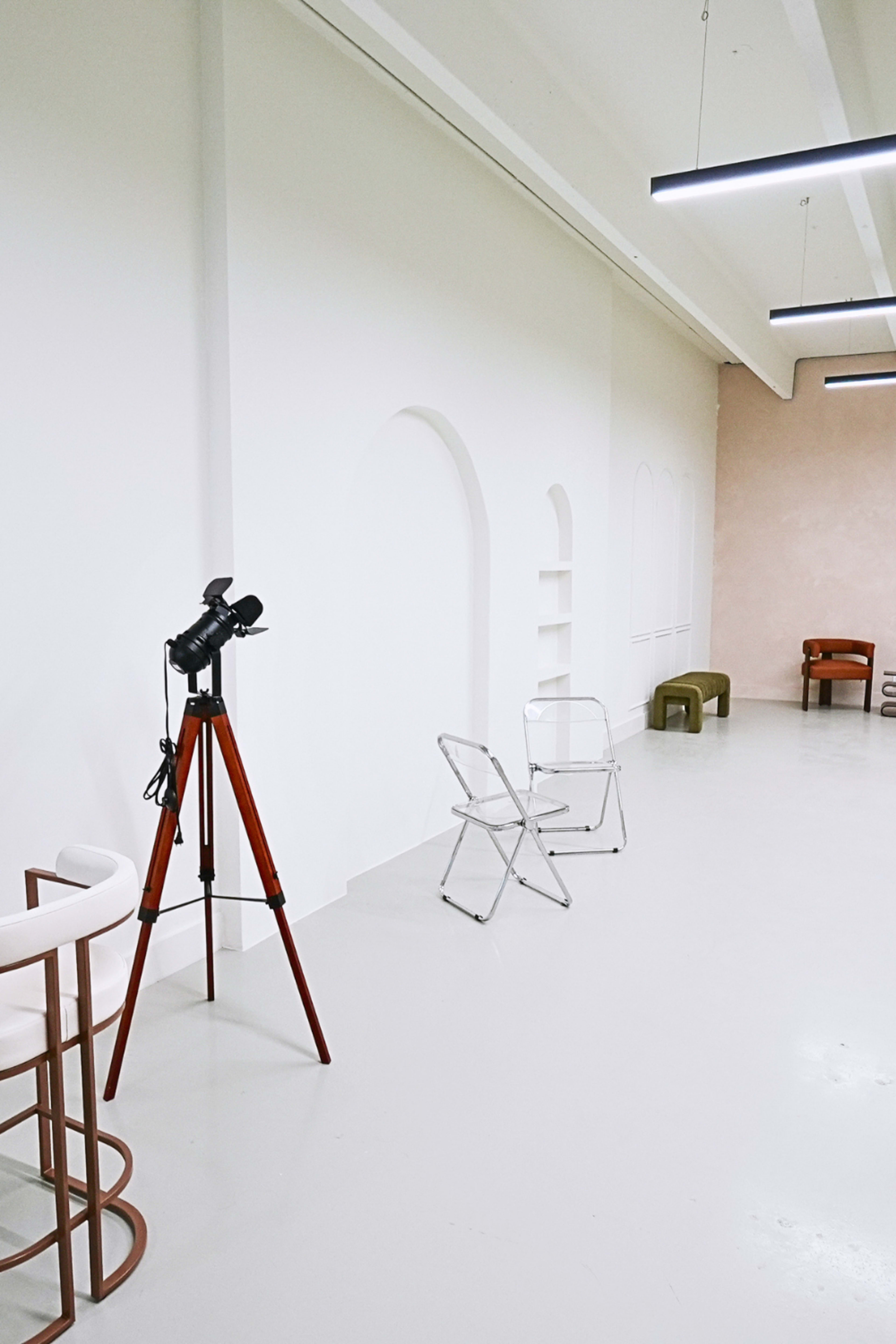 The image shows a minimalist interior space featuring a telescope on a wooden stand, two transparent chairs, and a few wooden furniture pieces against a neutral background.
