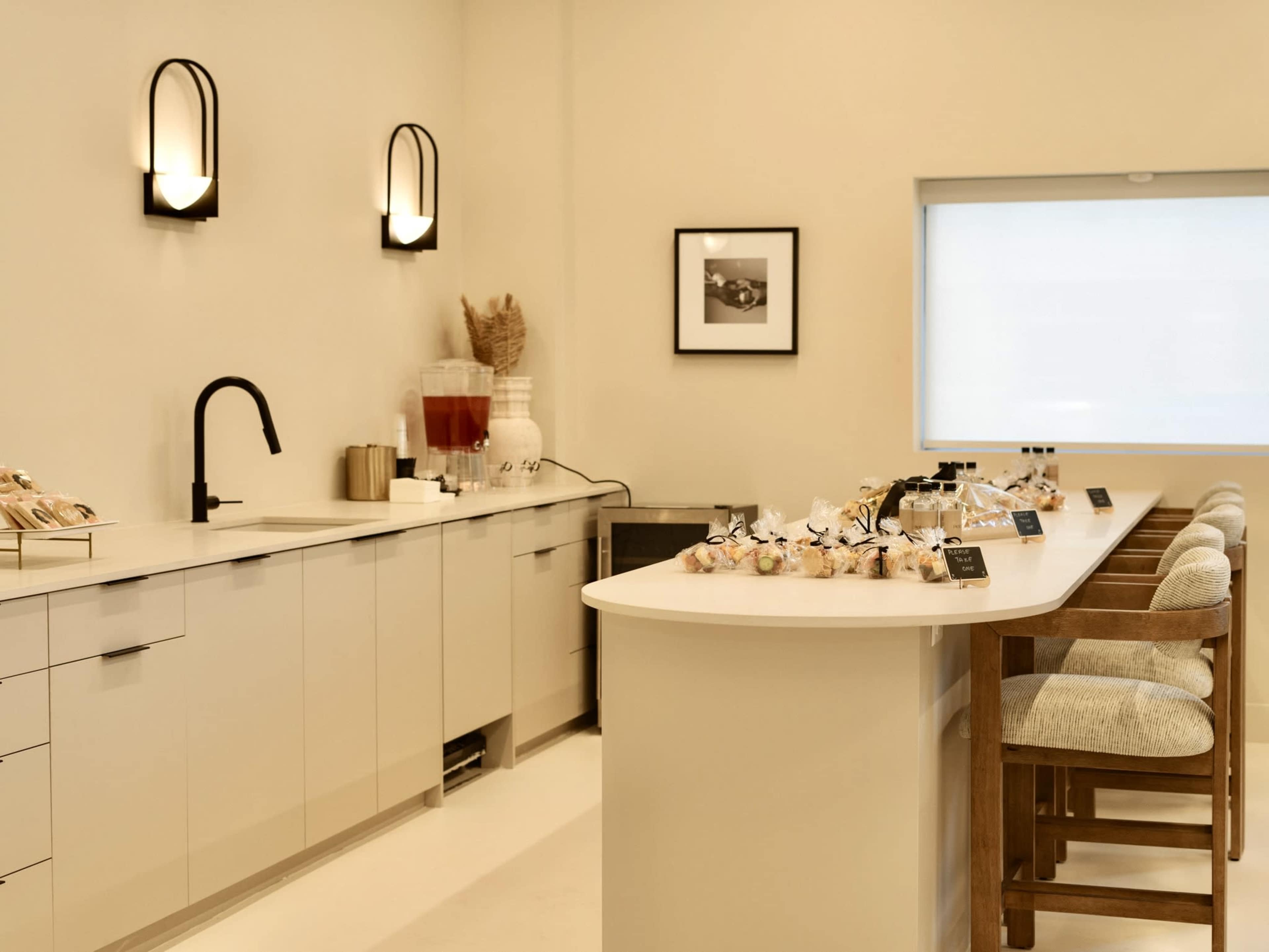 The image shows a modern kitchen space with a sleek counter, wooden stools, and a minimalist design featuring light-colored cabinetry and decorative items.