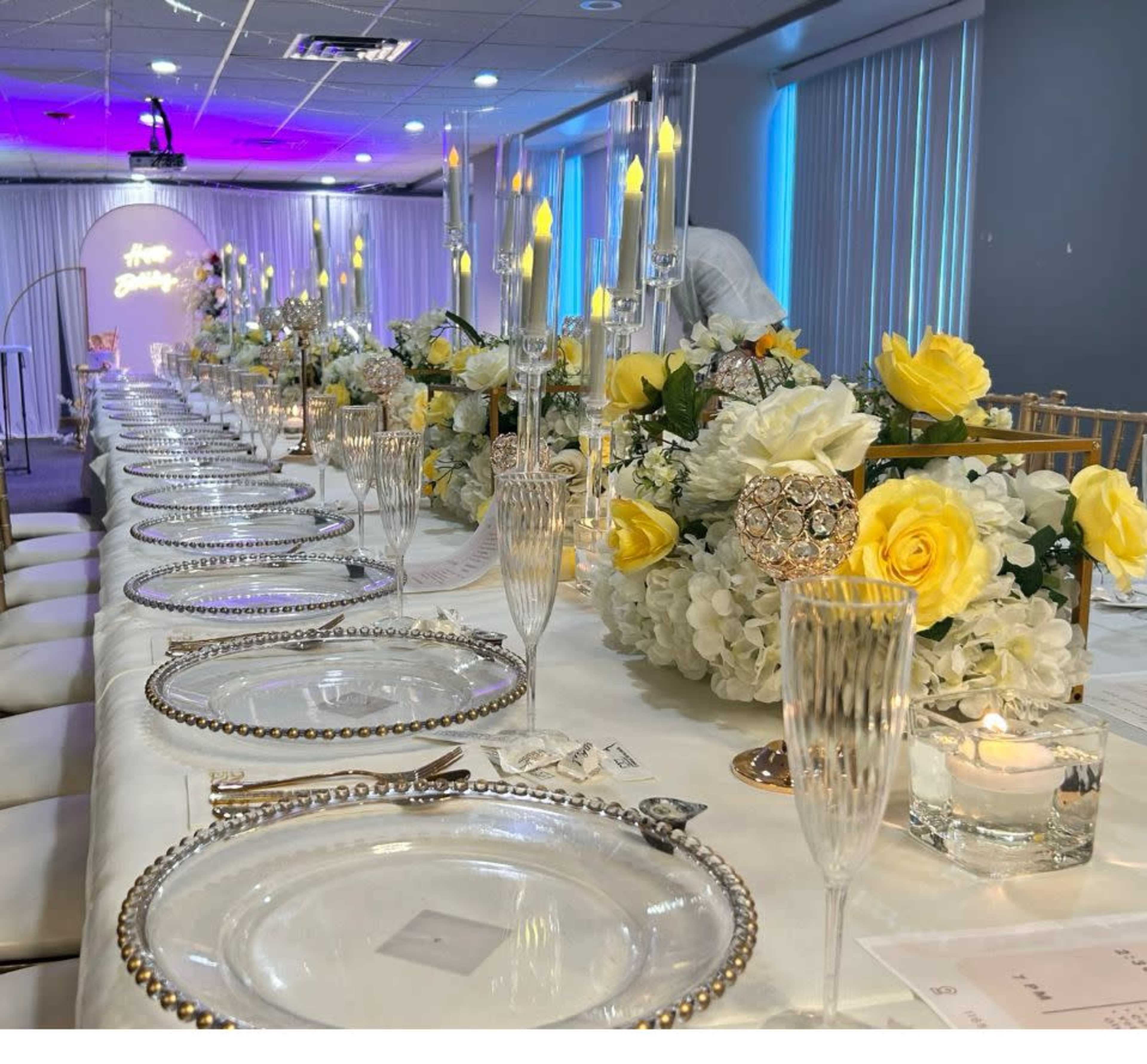 A long banquet table is elegantly arranged with decorative plates, crystal glassware, and floral centerpieces featuring yellow roses and white flowers.