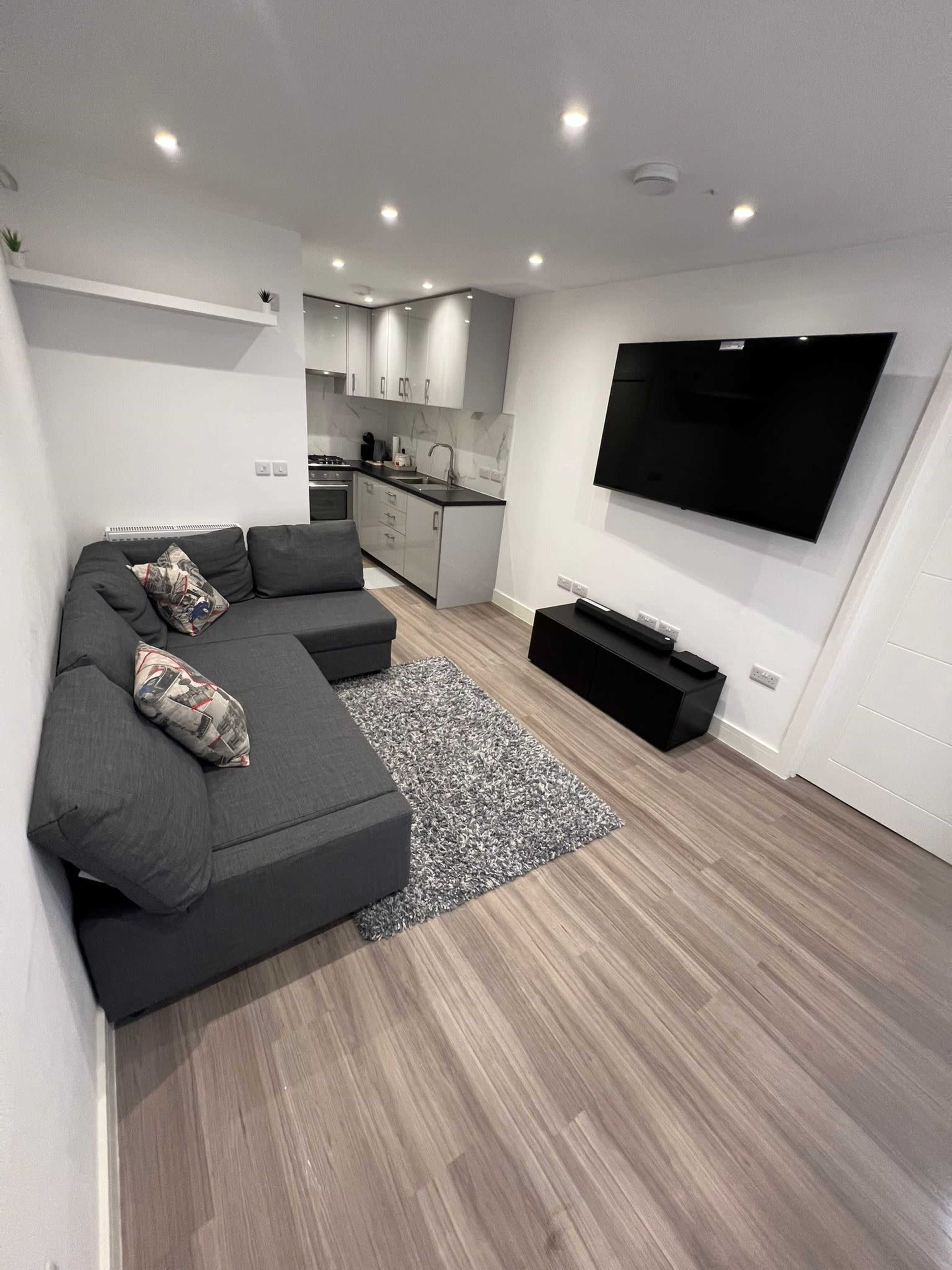 The image shows a modern living area with a gray sectional sofa, a wall-mounted television, and a kitchenette in the background.