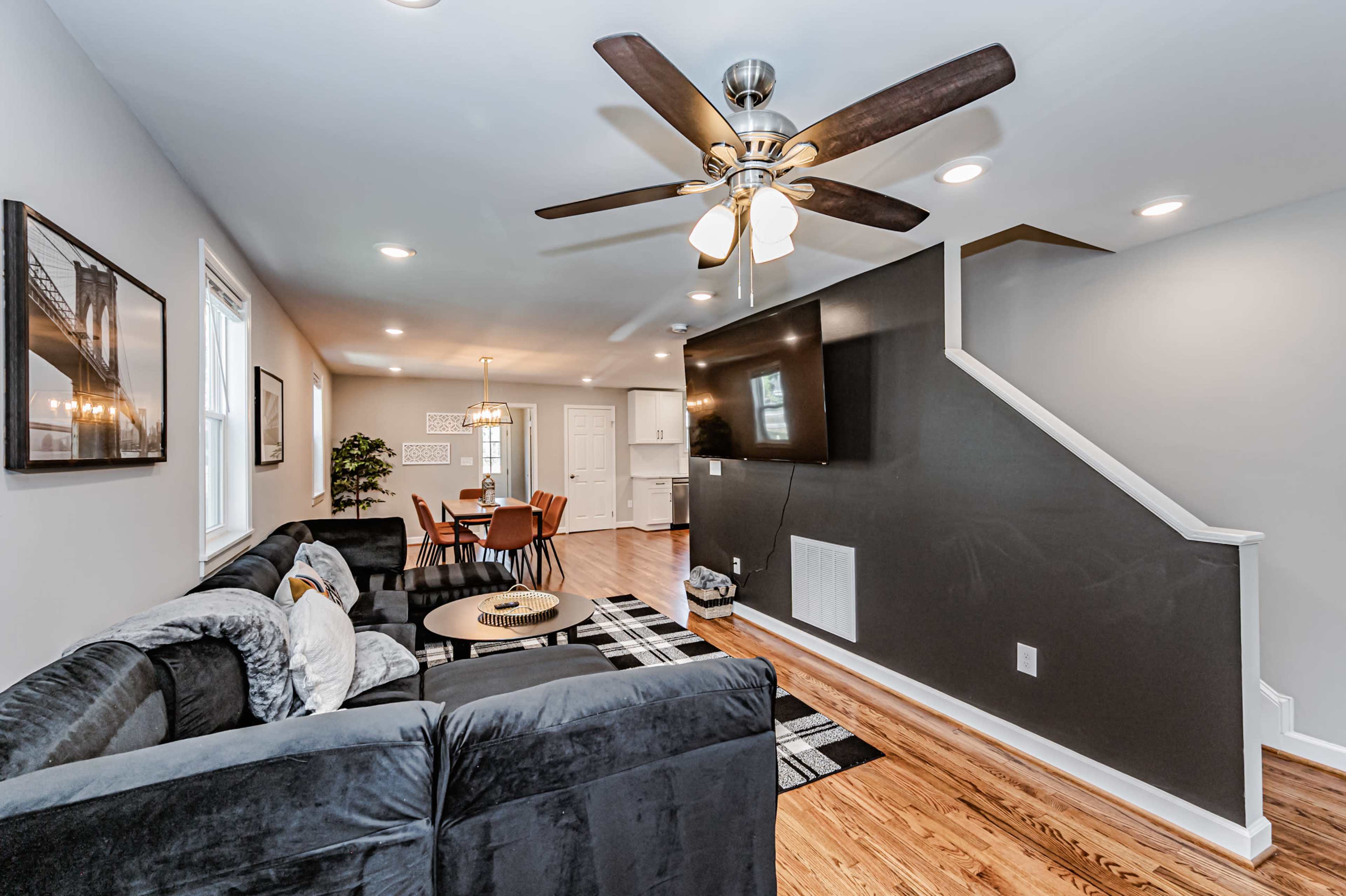 A living room with a dark sectional sofa, a ceiling fan, a mounted television on an accent wall, and a dining area in the background.