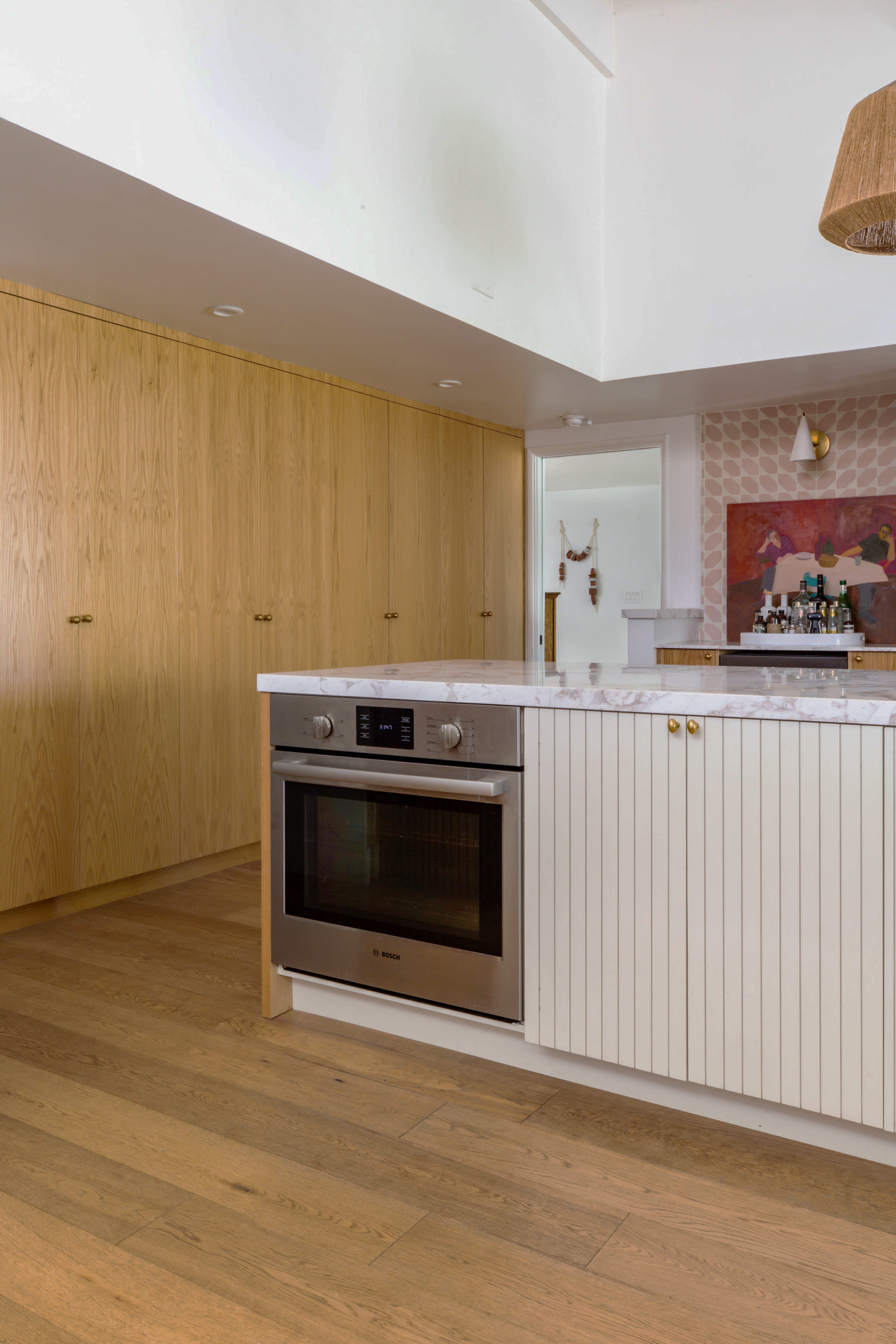 The image shows a modern kitchen featuring light wood cabinetry, a white countertop with an integrated oven, and a decorative wall with muted art.
