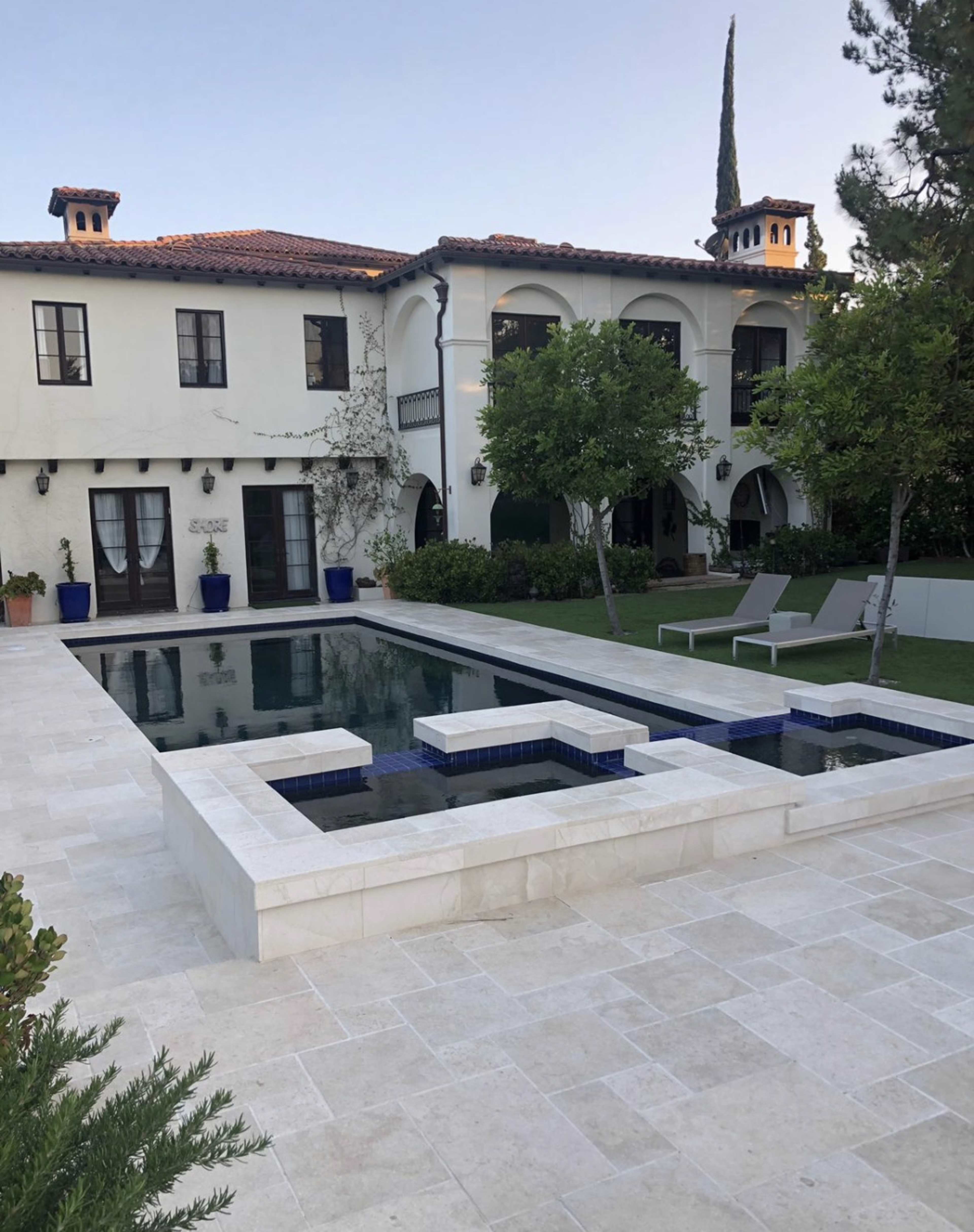 The image shows a modern home with a two-story facade overlooking a rectangular pool surrounded by a stone patio and landscaped yard.