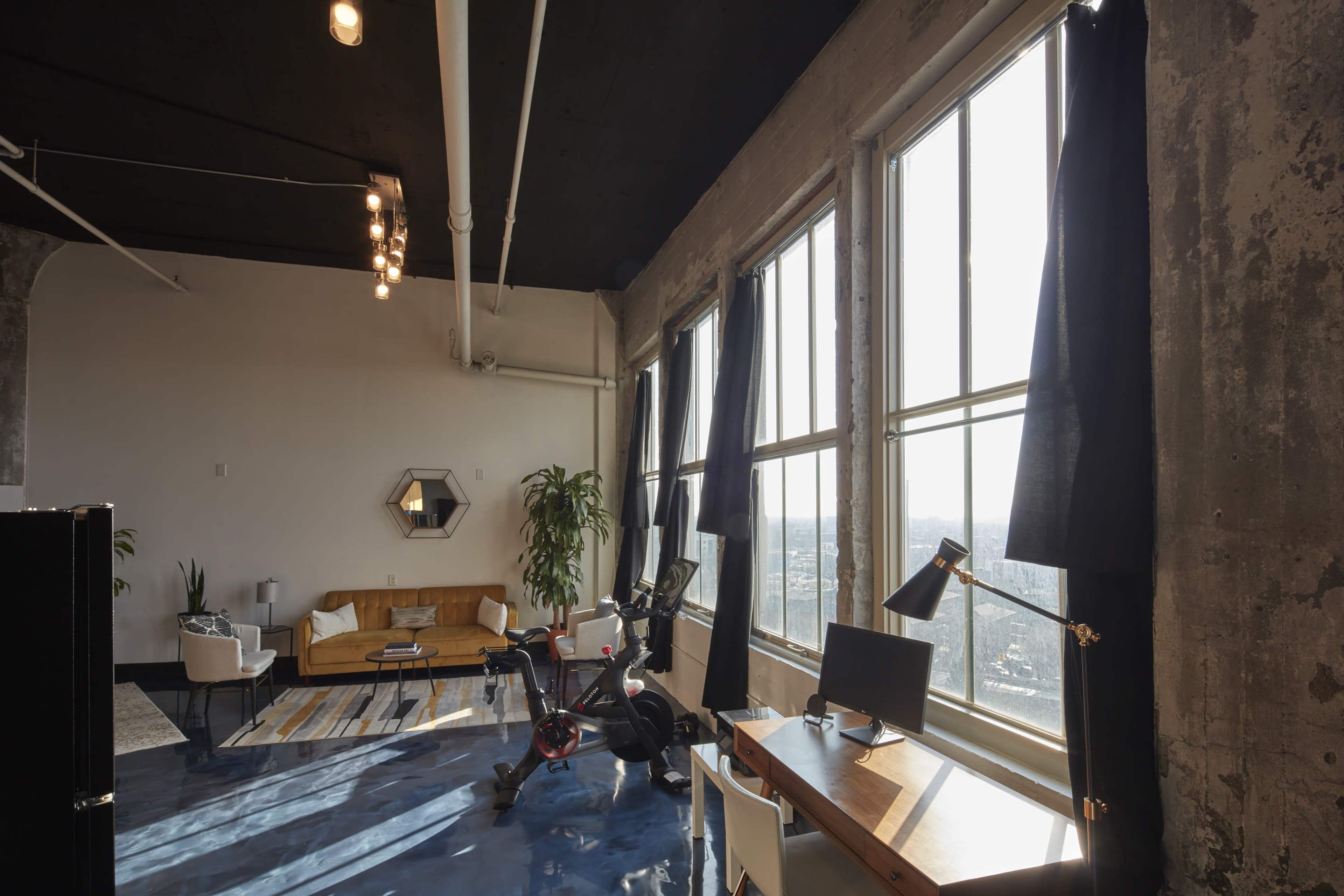 A spacious industrial-style room features large windows, a yellow sofa, a bike, a desk with a monitor, and a few potted plants.
