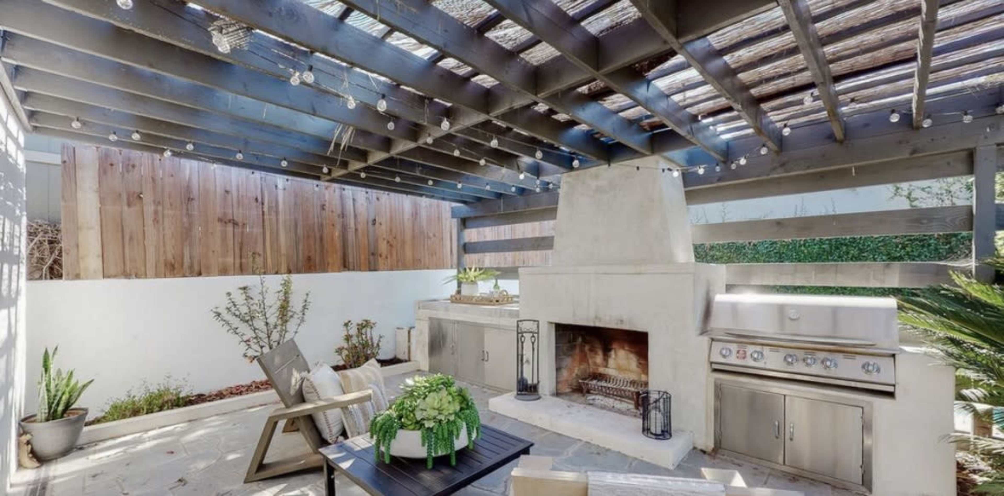 The image shows an outdoor kitchen area with a fireplace, grill, and seating, covered by a wooden pergola and decorated with string lights.