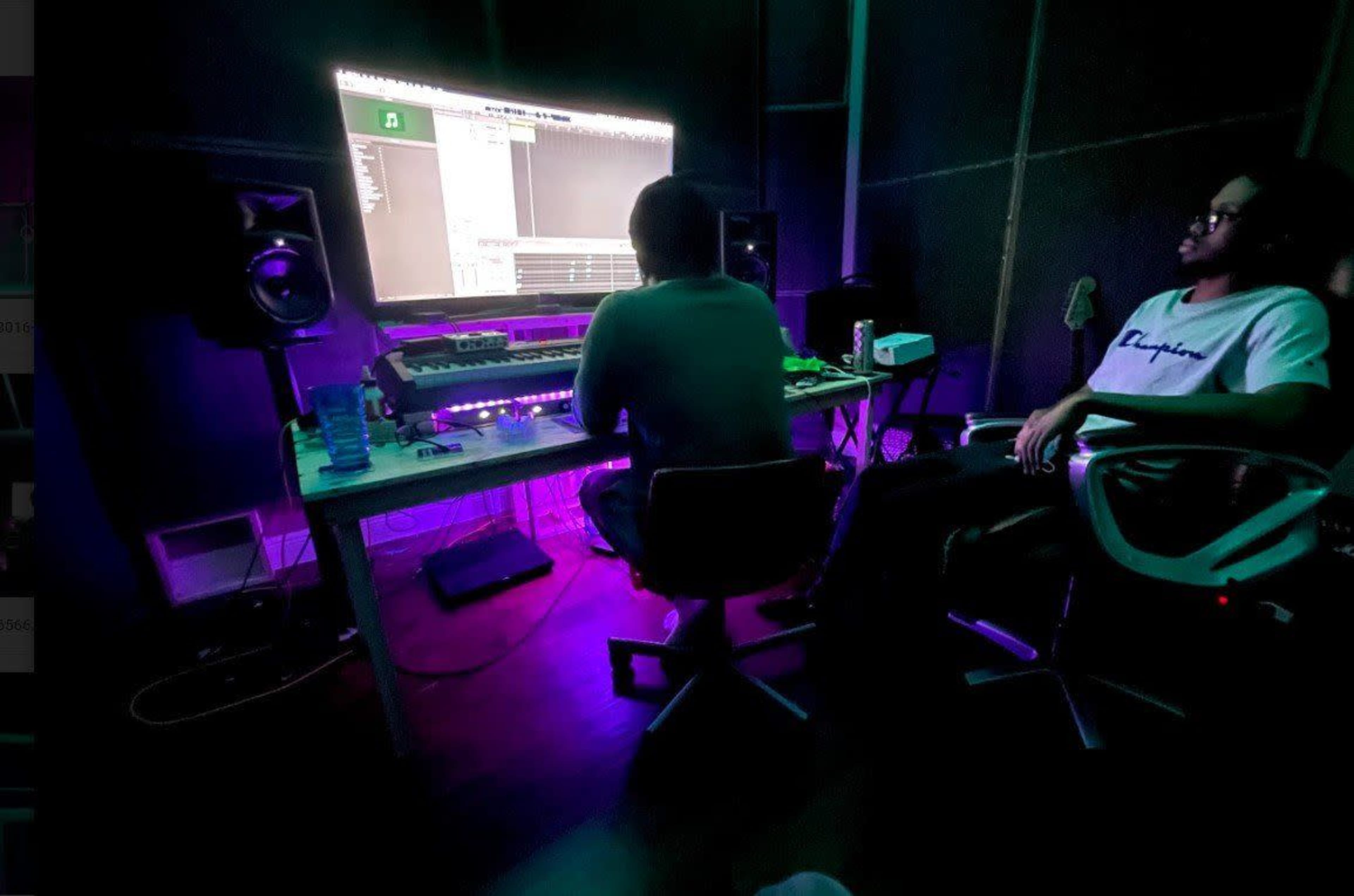 Two individuals are seated in a dimly lit room with a large computer monitor displaying music software, surrounded by audio equipment.