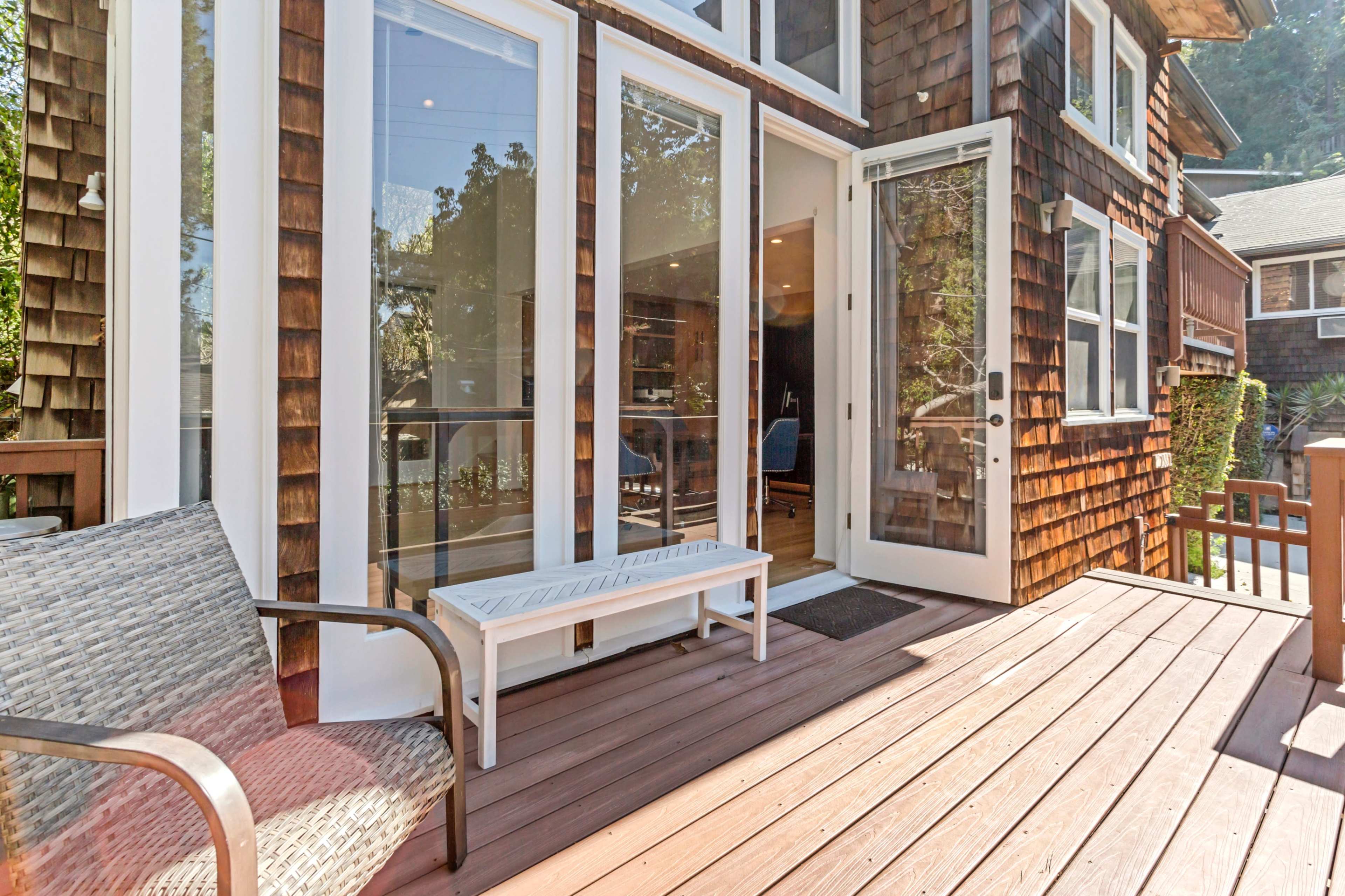 The image shows a wooden deck with a bench and a doorway leading into a well-lit room of a house with shingle siding.