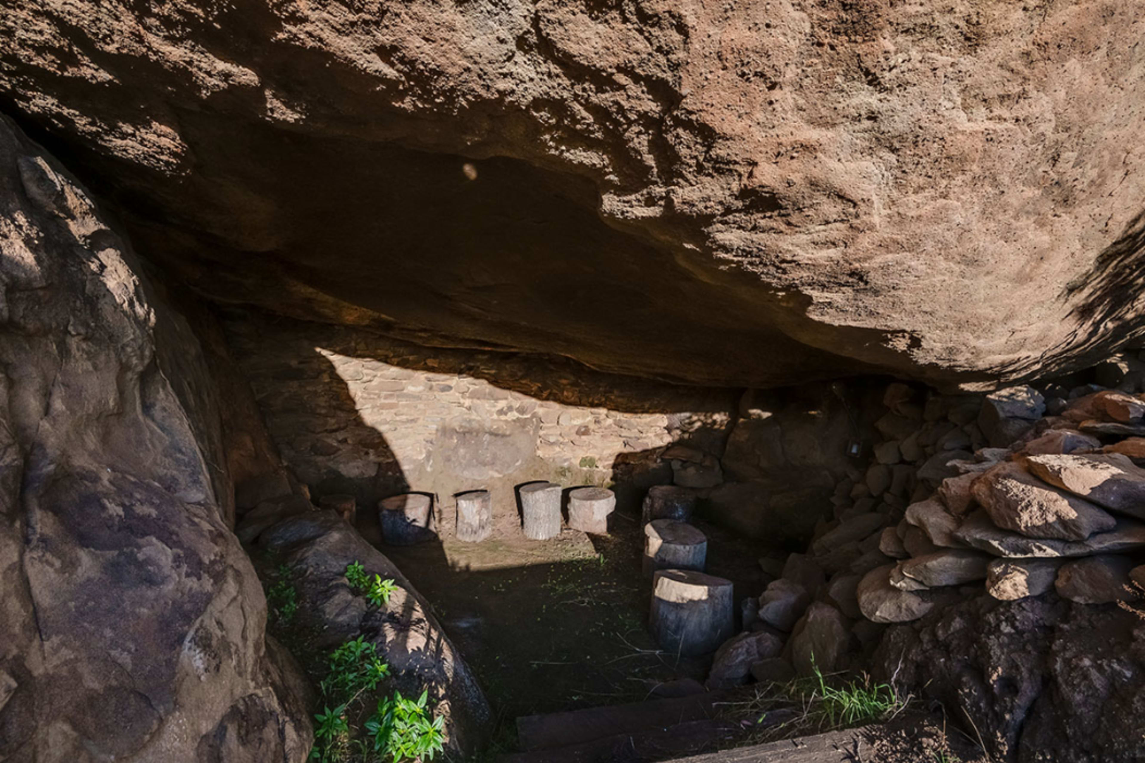 A small, natural cave features a stone floor and circular seating arranged around a central area, set against a backdrop of large rock formations.