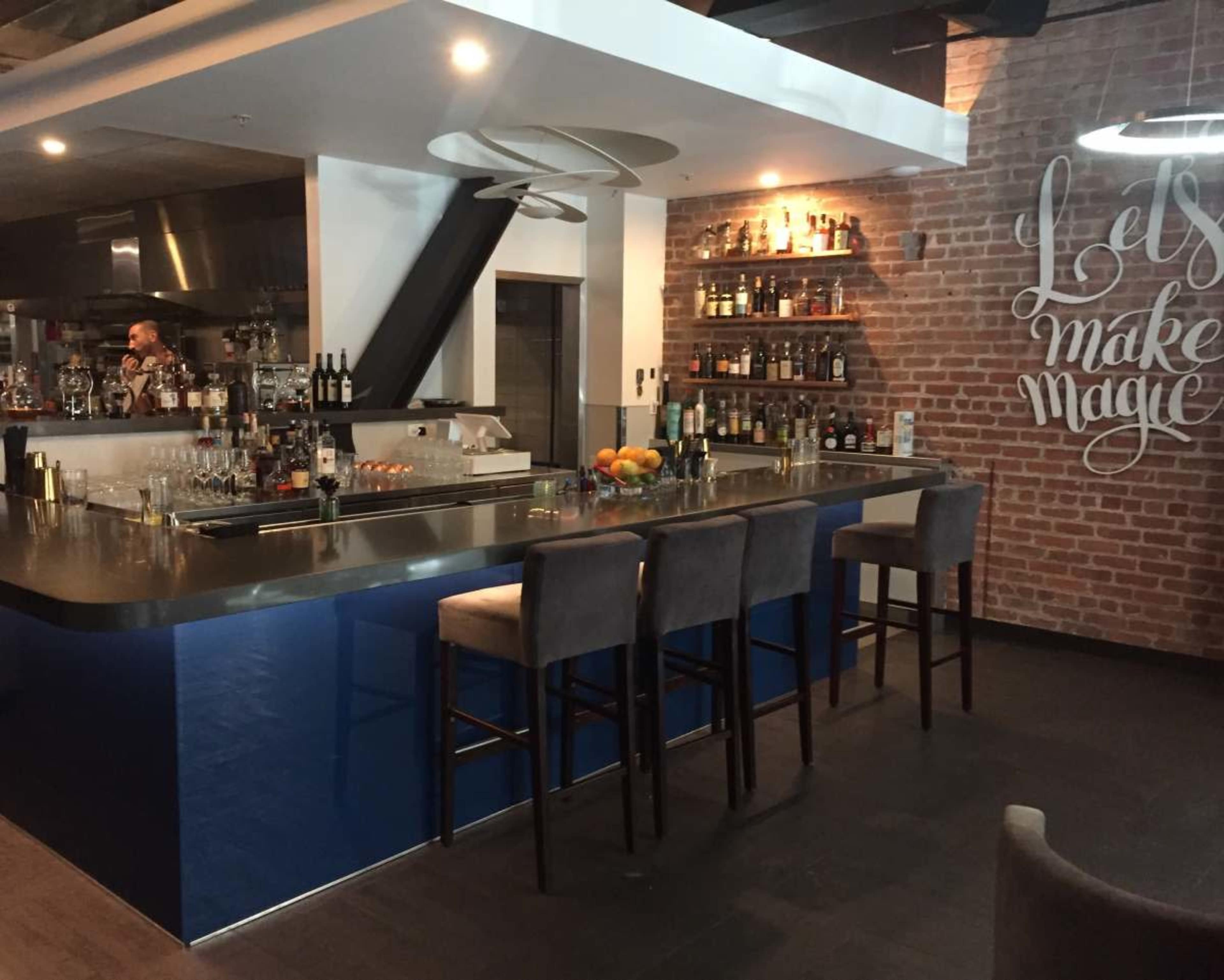 The image shows a modern bar area with a sleek counter, a variety of bottles displayed on shelves, and high stools positioned along the bar.