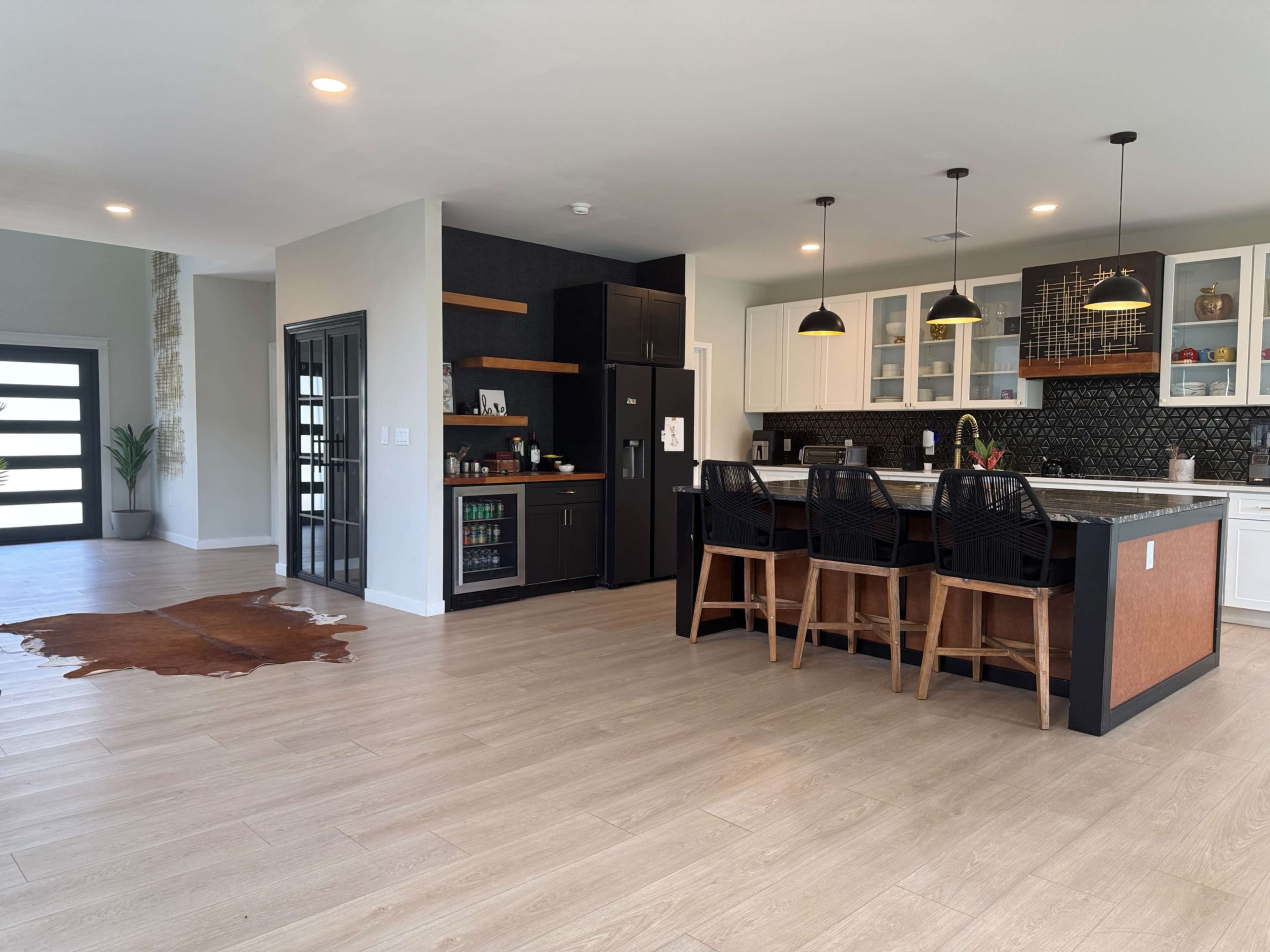 The image depicts a modern open-concept kitchen and dining area featuring a large island with bar stools, a dark refrigerator, and a living room space with a cowhide rug on the wooden floor.