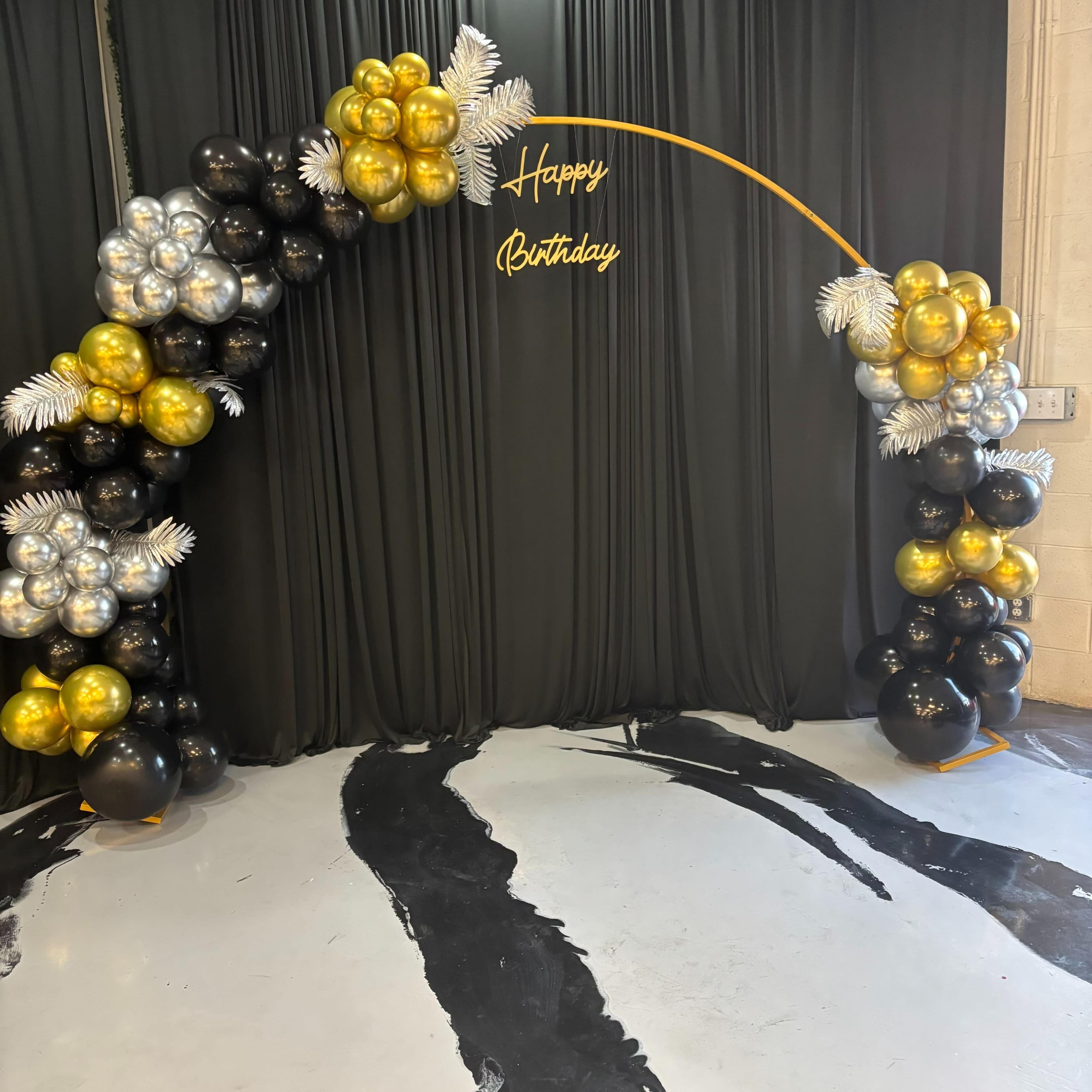 A decorative balloon arch in black, gold, and silver is set against a black backdrop, with the words "Happy Birthday" displayed in the center.