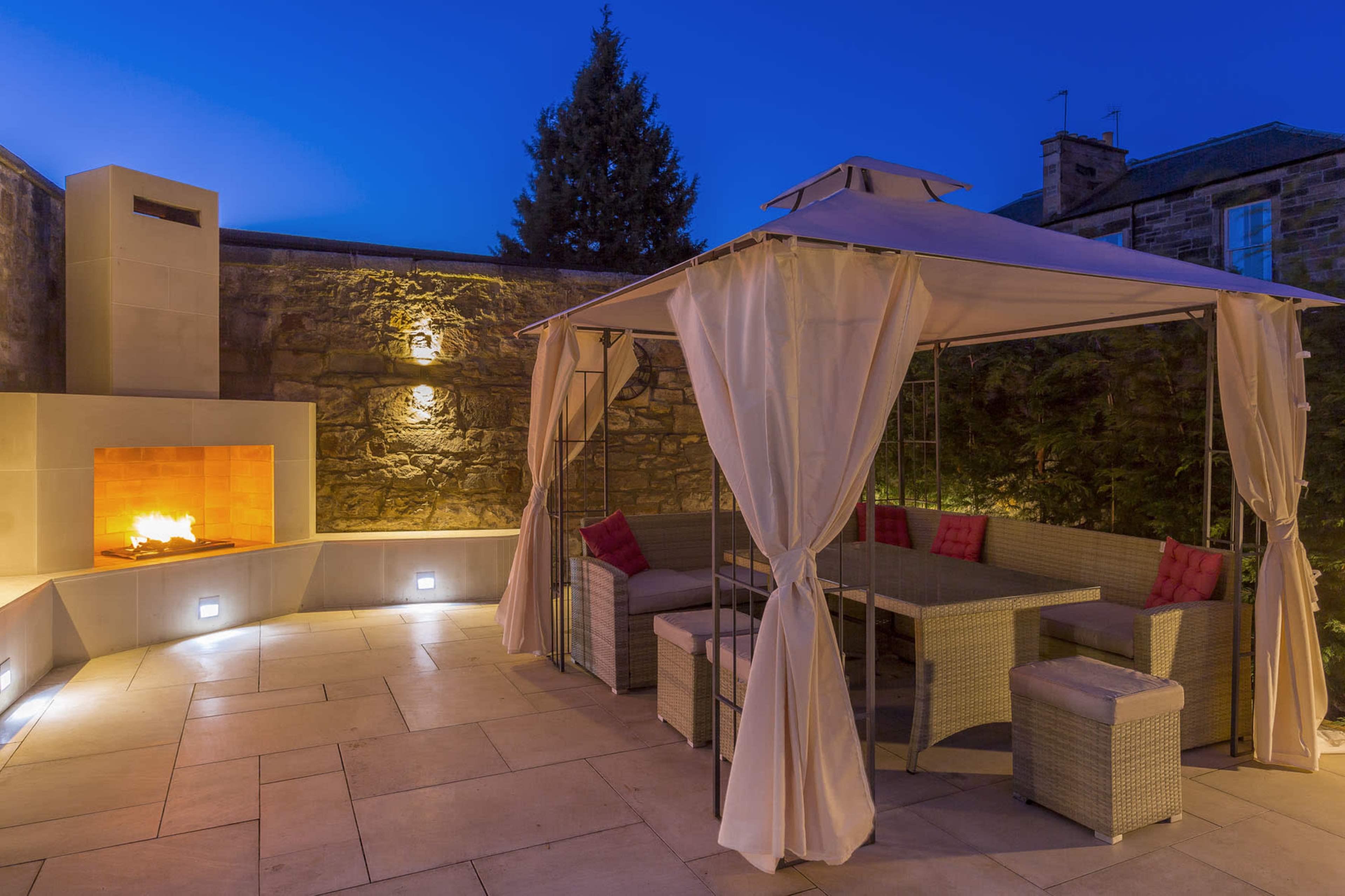 A patio area featuring a gazebo with draped curtains, a seating arrangement, and an outdoor fireplace, illuminated at dusk.