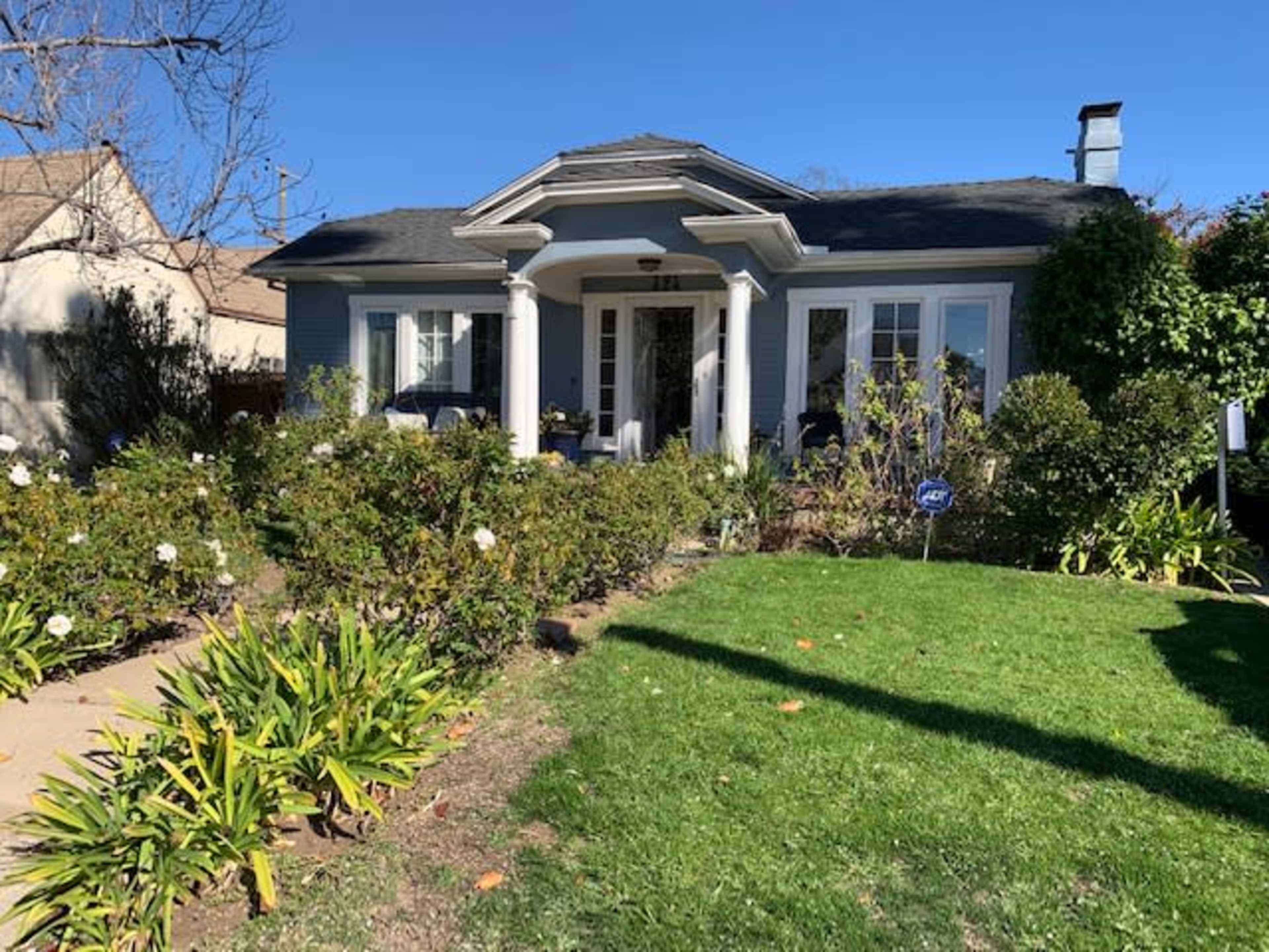 A single-story house with a covered porch, surrounded by green shrubs and flowers, sits on a well-maintained lawn.