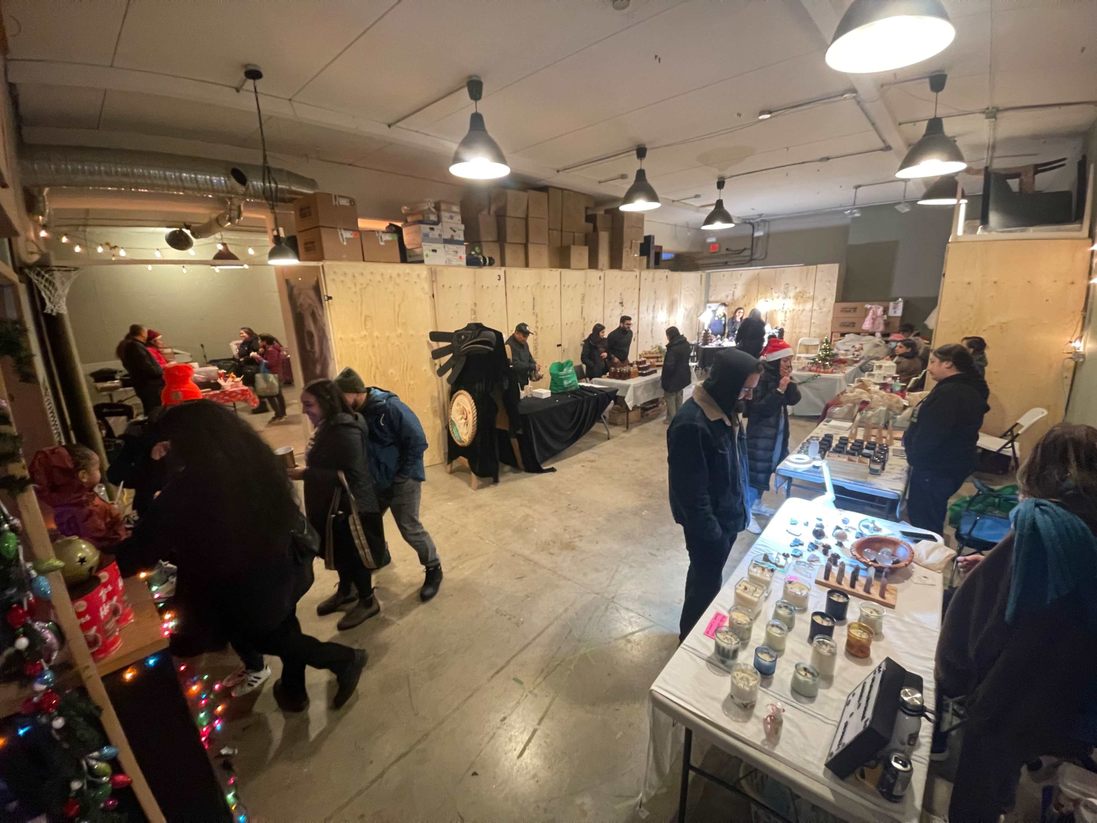 A crowded indoor market features various vendor tables displaying goods, with shoppers browsing among the stalls.