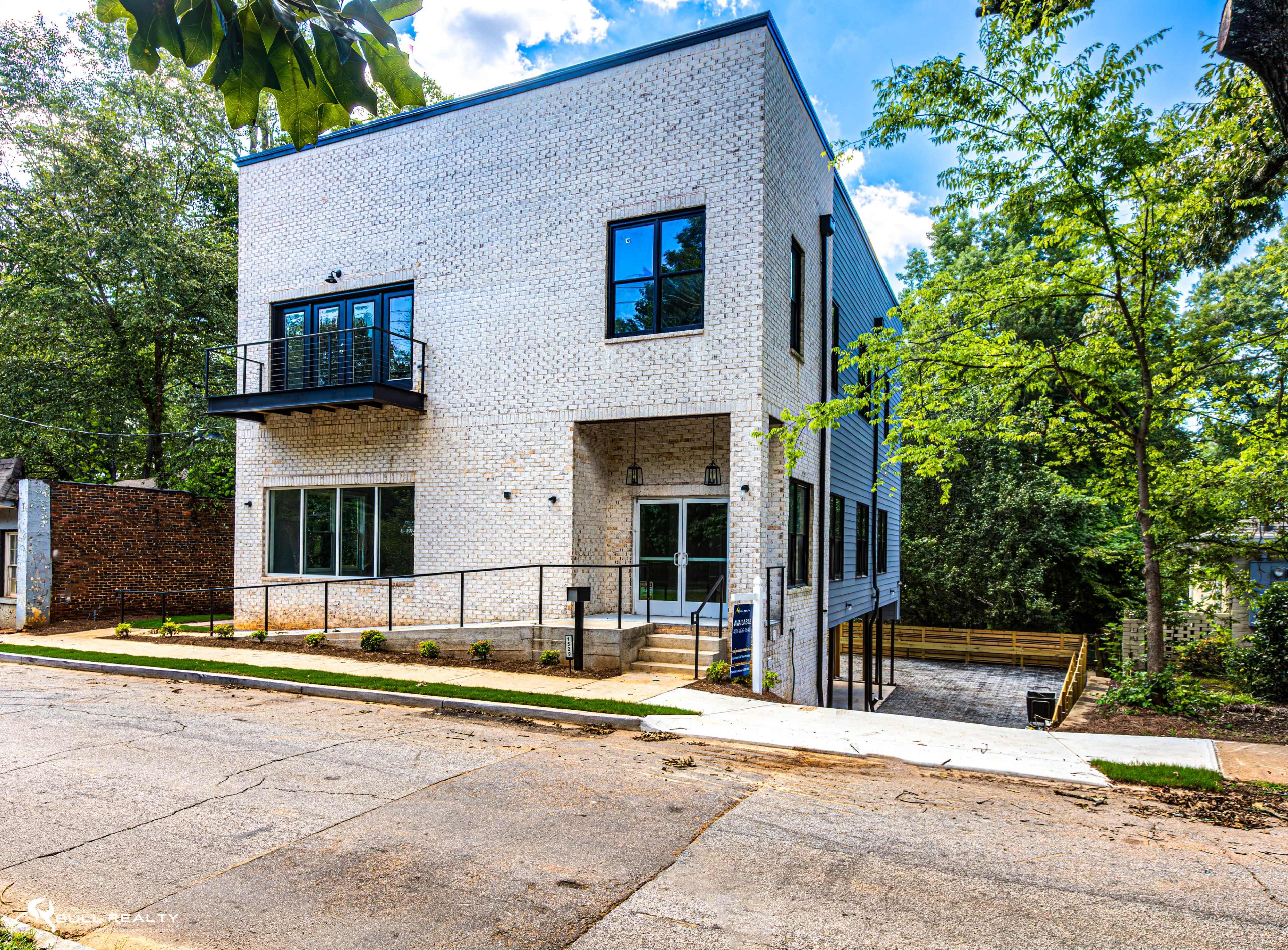 The image shows a modern two-story building with white brick exterior and large windows, situated on a sloped street surrounded by greenery.