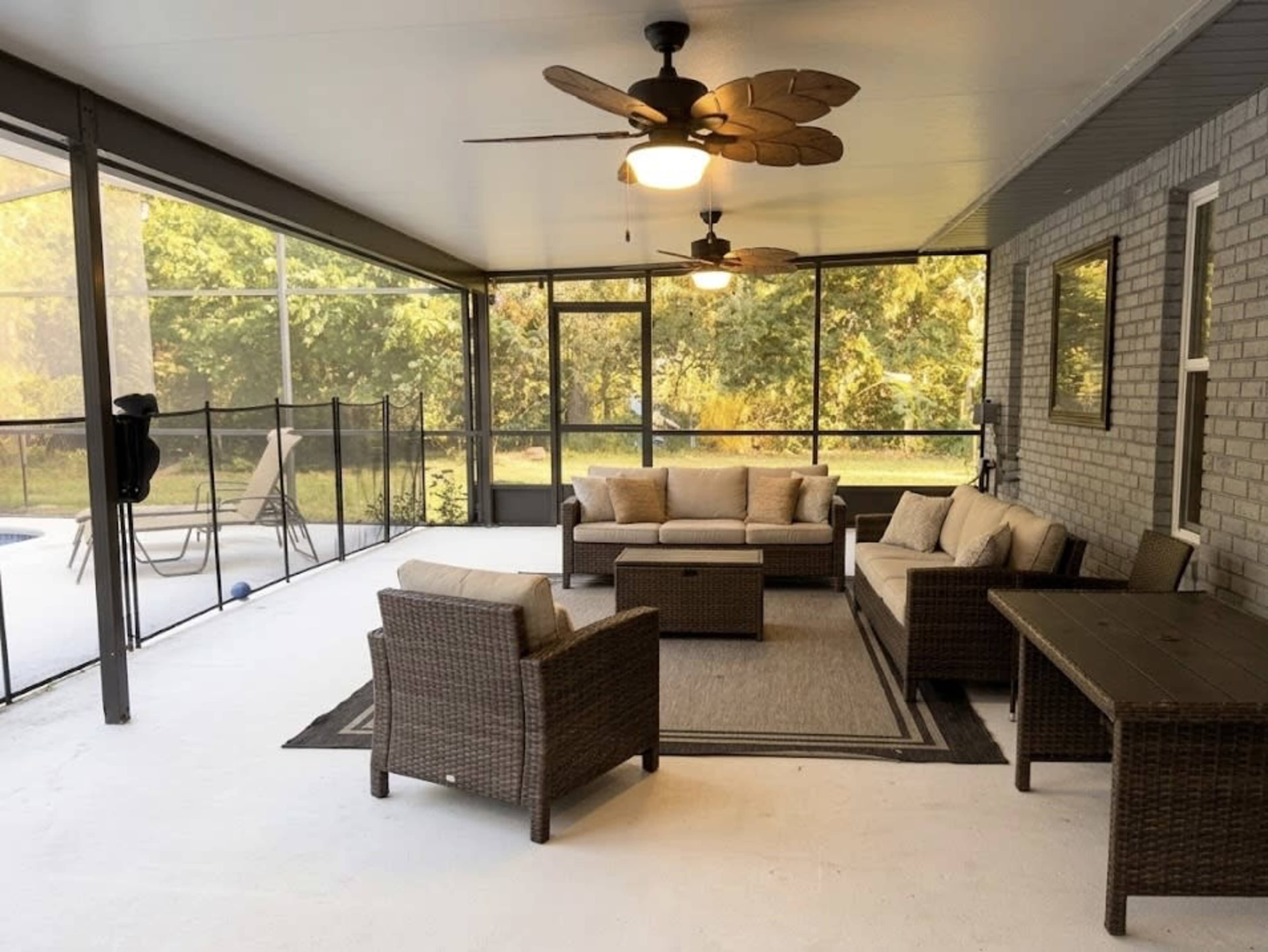 The image shows a screened-in porch furnished with a wicker seating arrangement, a coffee table, and ceiling fans, set against a backdrop of trees.