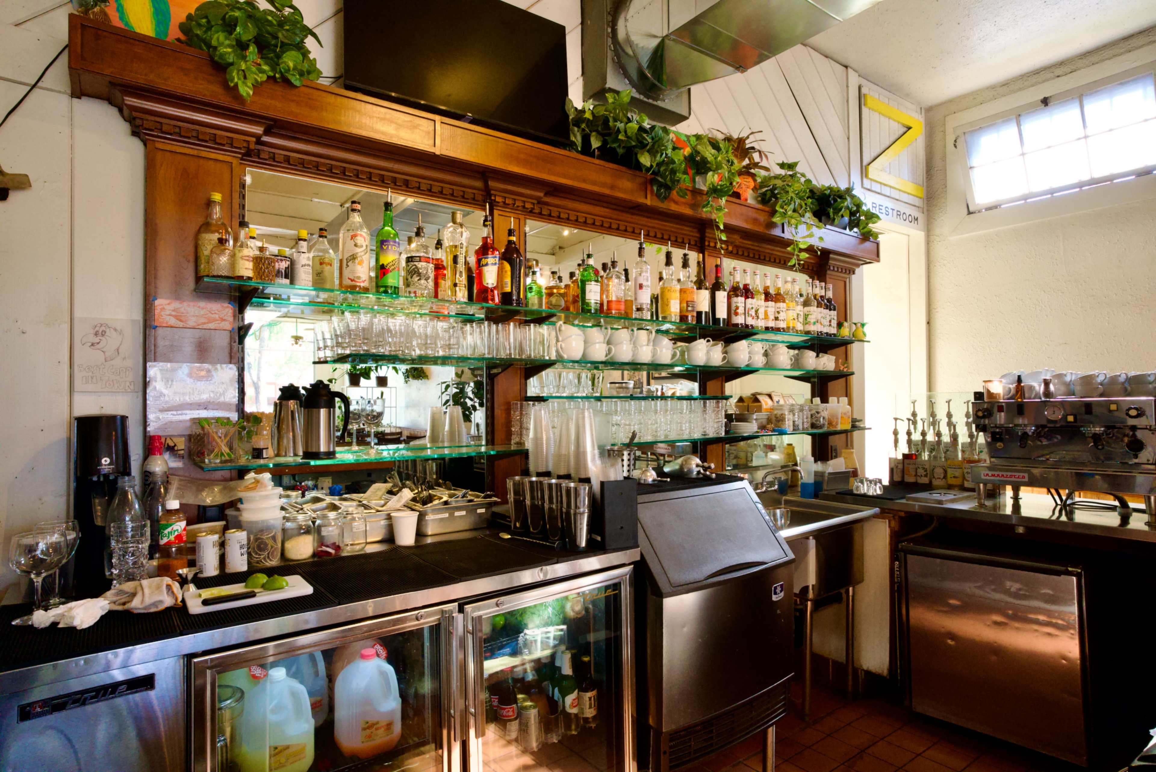 The image shows a well-organized bar area with a large wooden shelf displaying various bottles of liquor, glassware, and a coffee machine.