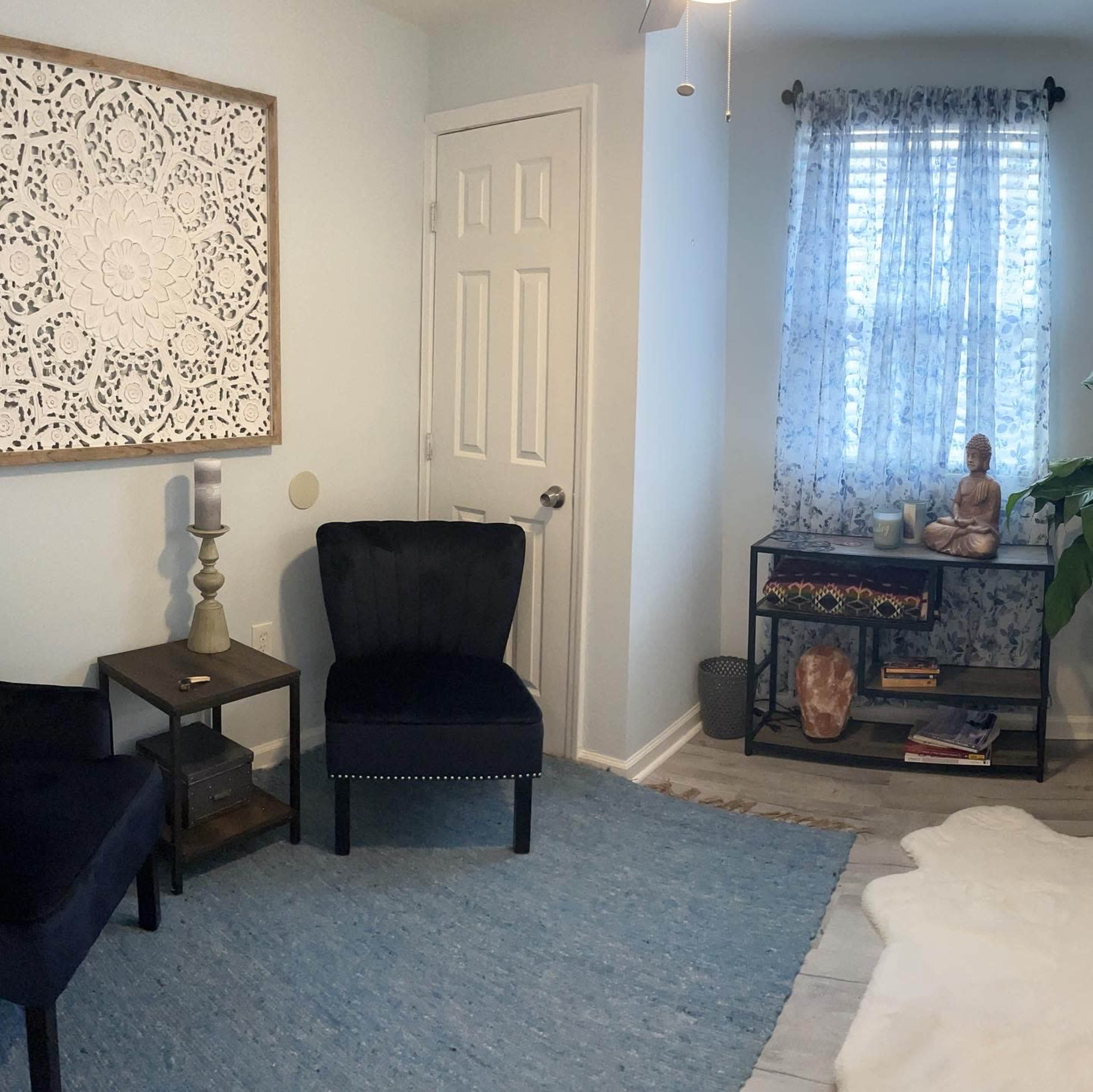 The room features two black chairs, a patterned wall hanging, a window with sheer curtains, and a small plant beside a shelf with decorative items.