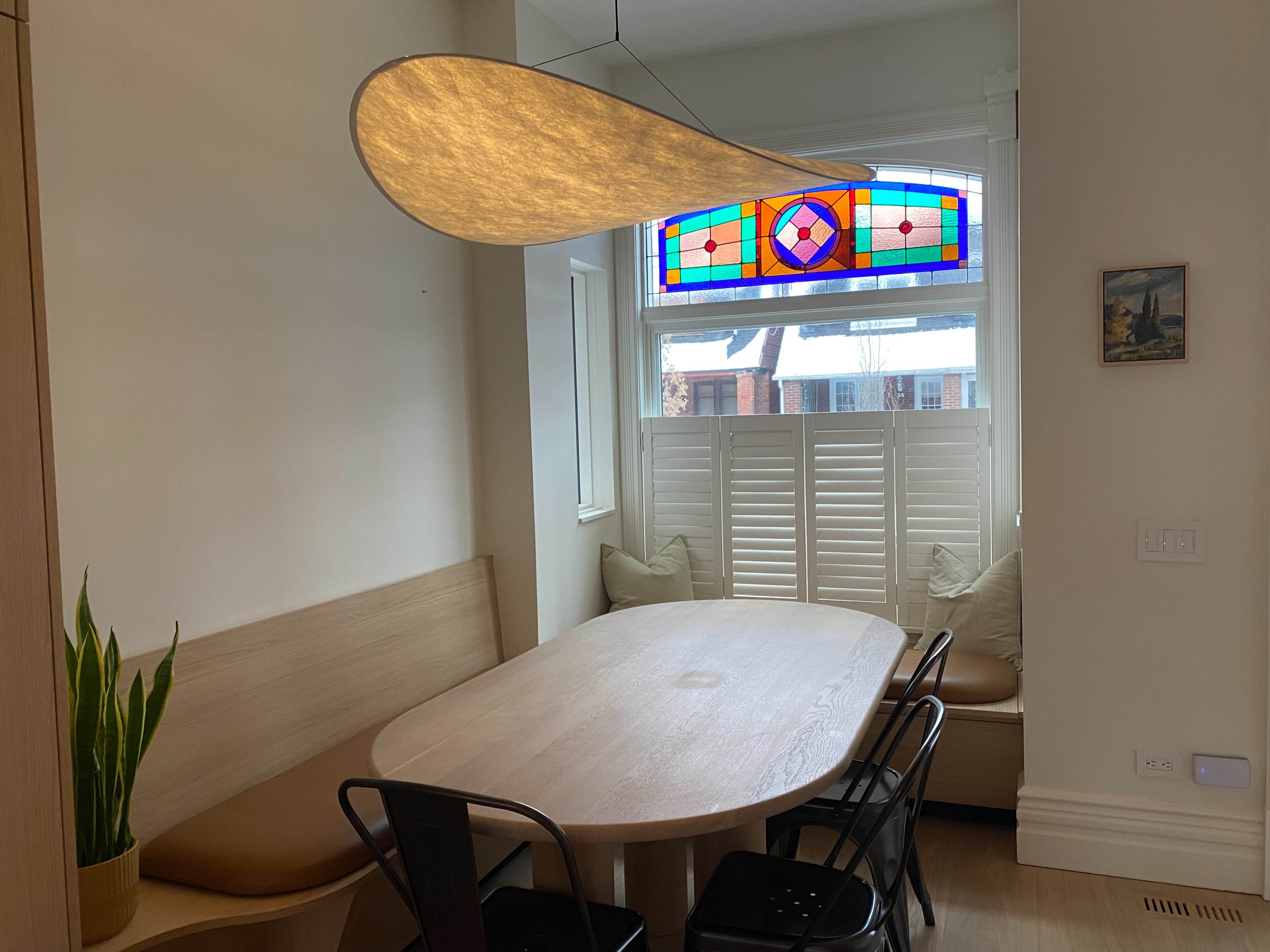 The image shows a dining area featuring a large wooden table surrounded by black chairs, with a light fixture overhead and a stained glass window that illuminates the space.