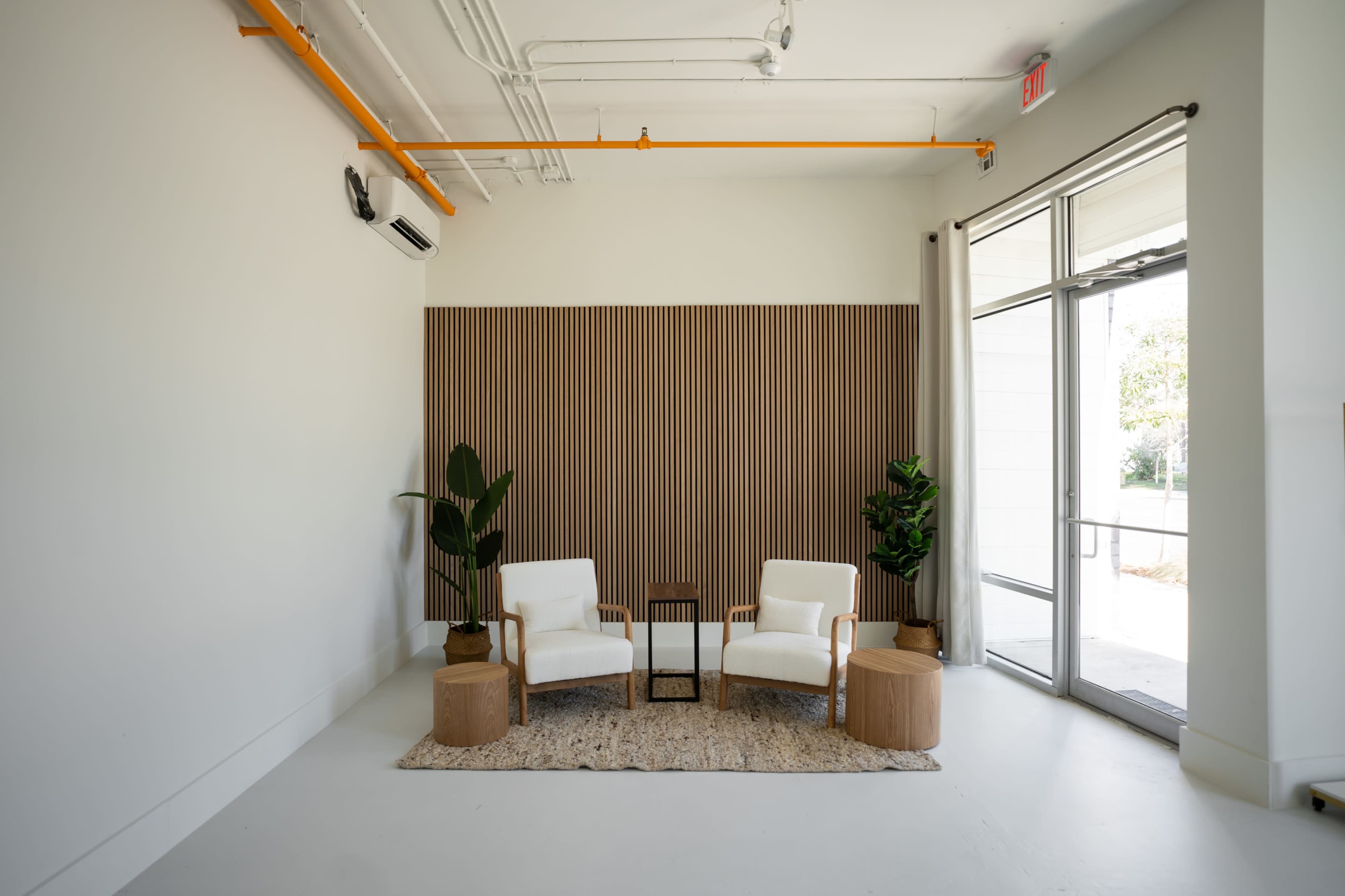 A minimalist interior space with two white chairs, a small table, and plants beside a wood panel backdrop.