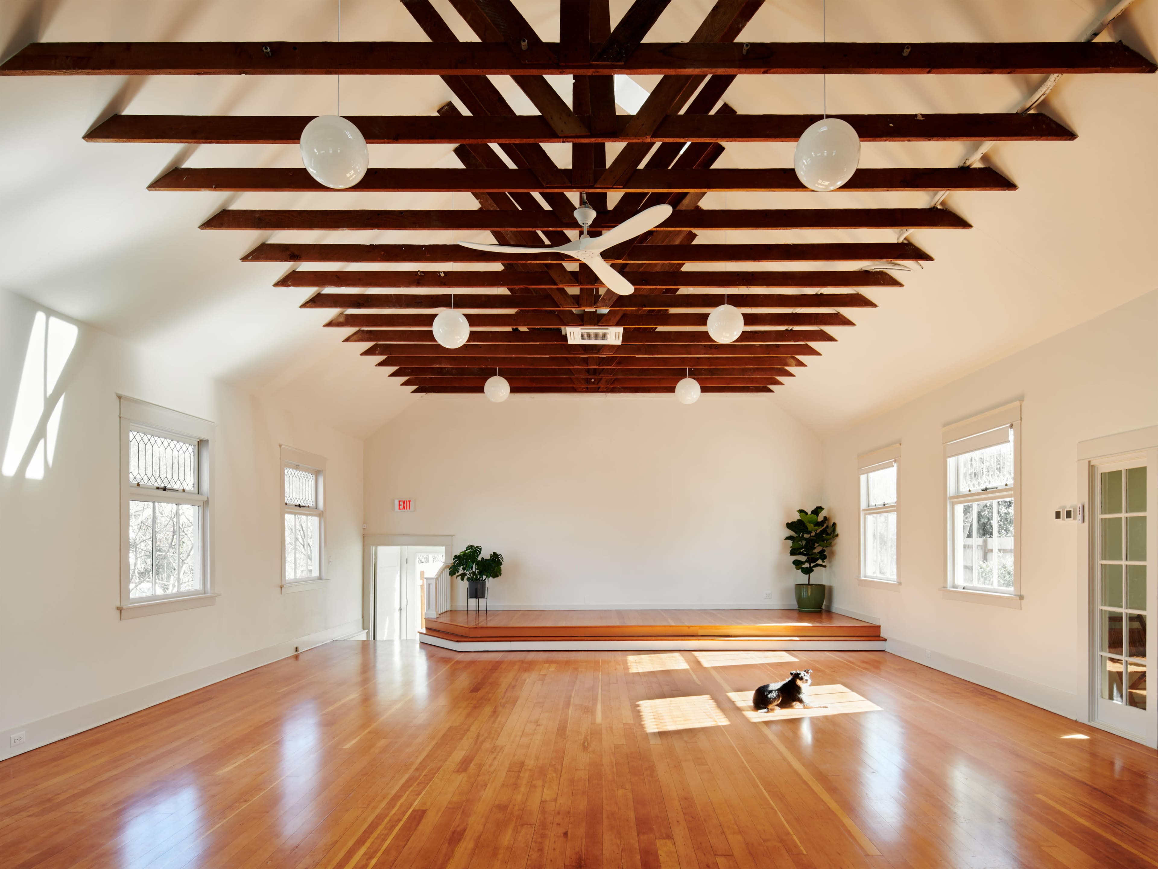 A spacious, empty room with wooden beams, large windows, and a wooden stage at one end.