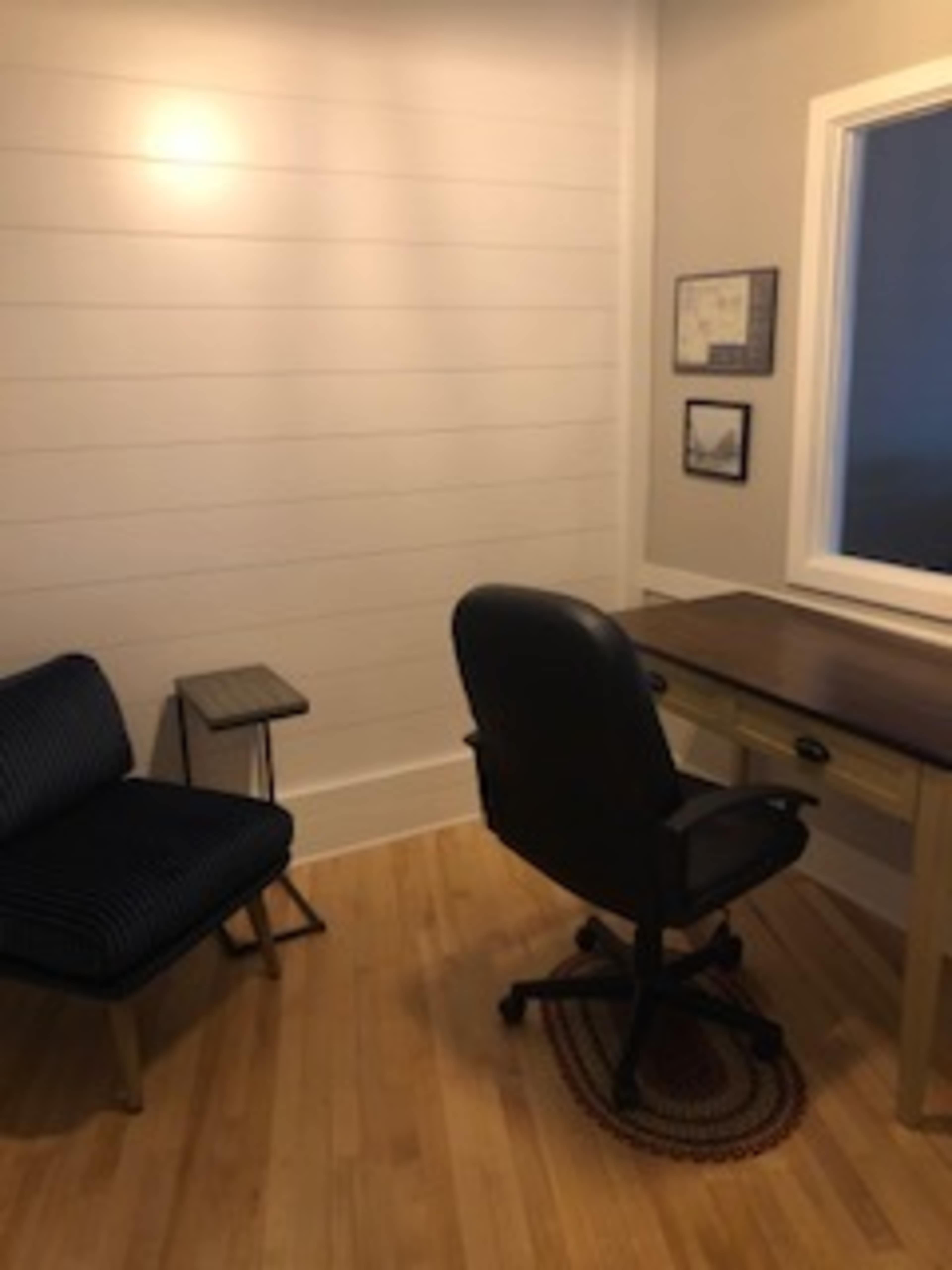 A small, minimalist office space with a wooden desk and a black office chair, accompanied by a blue-striped armchair and a small side table.