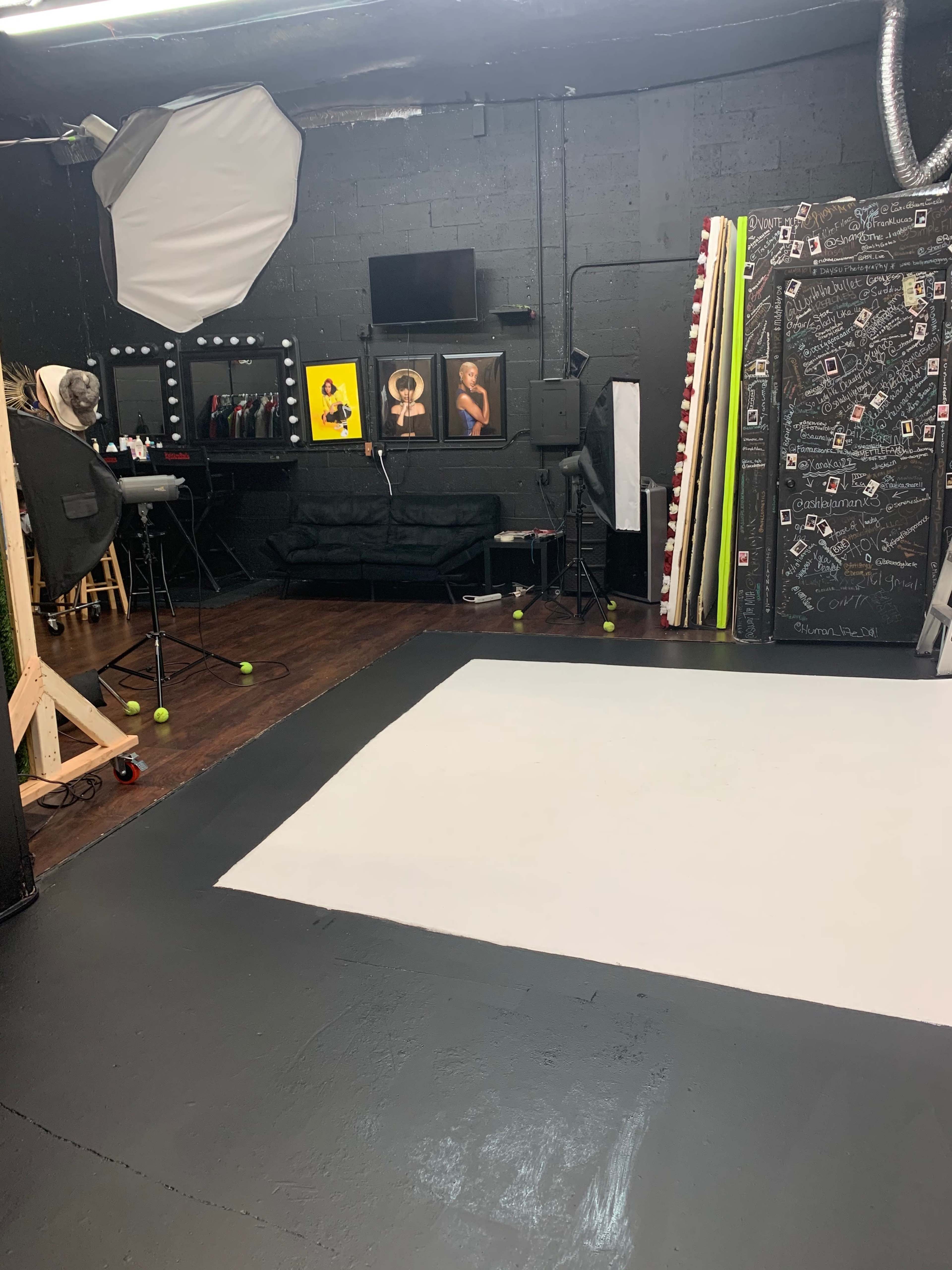 A photography studio with a gray floor, a backdrop setup, framed pictures on the wall, and various equipment arranged in a corner.
