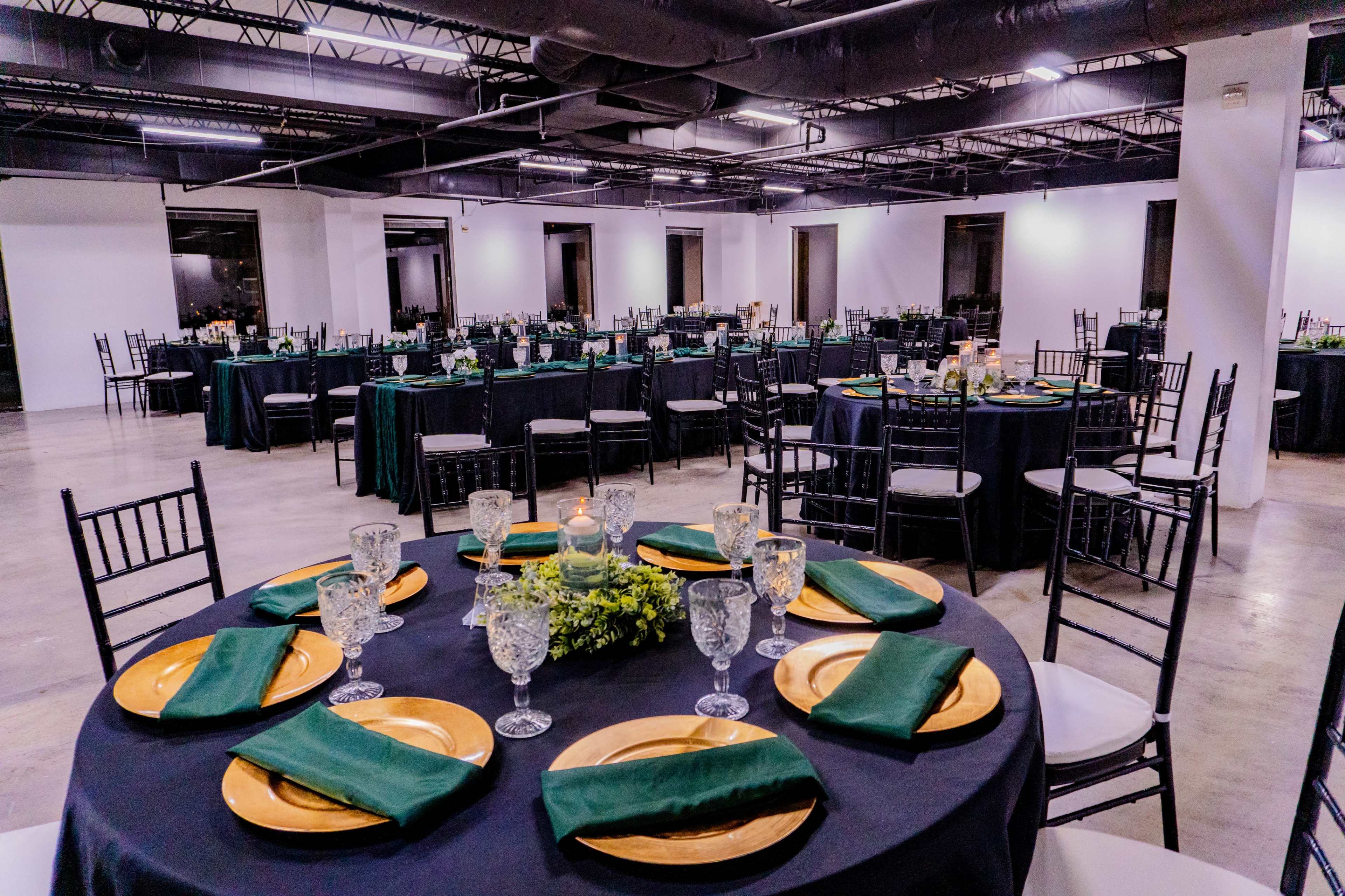 A spacious event room features round tables set with dark tablecloths, gold chargers, green napkins, and glassware, surrounded by additional tables and chairs.