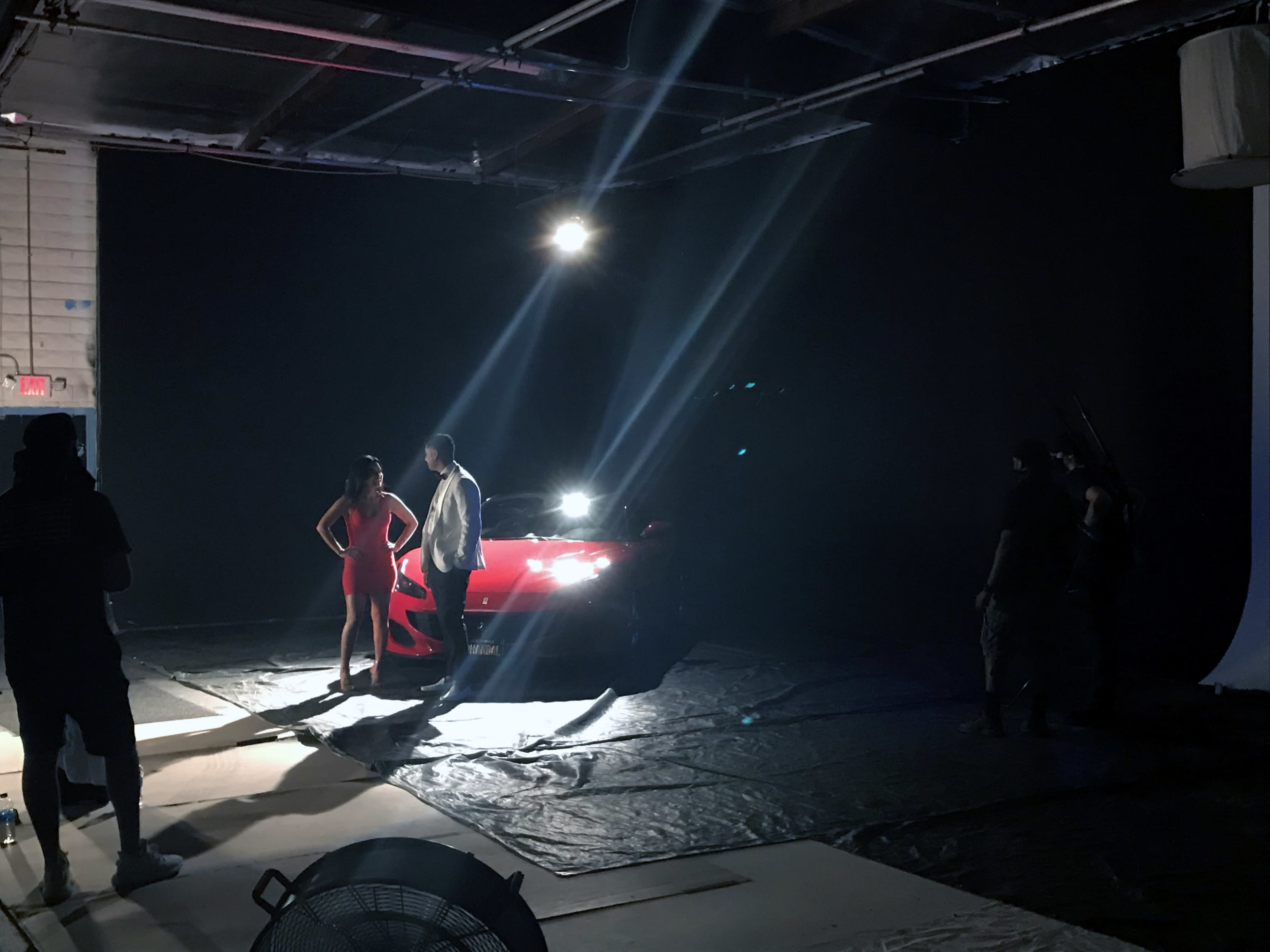 A red car is positioned in a dimly lit studio while two individuals pose in front of it, with crew members operating cameras and lights in the background.