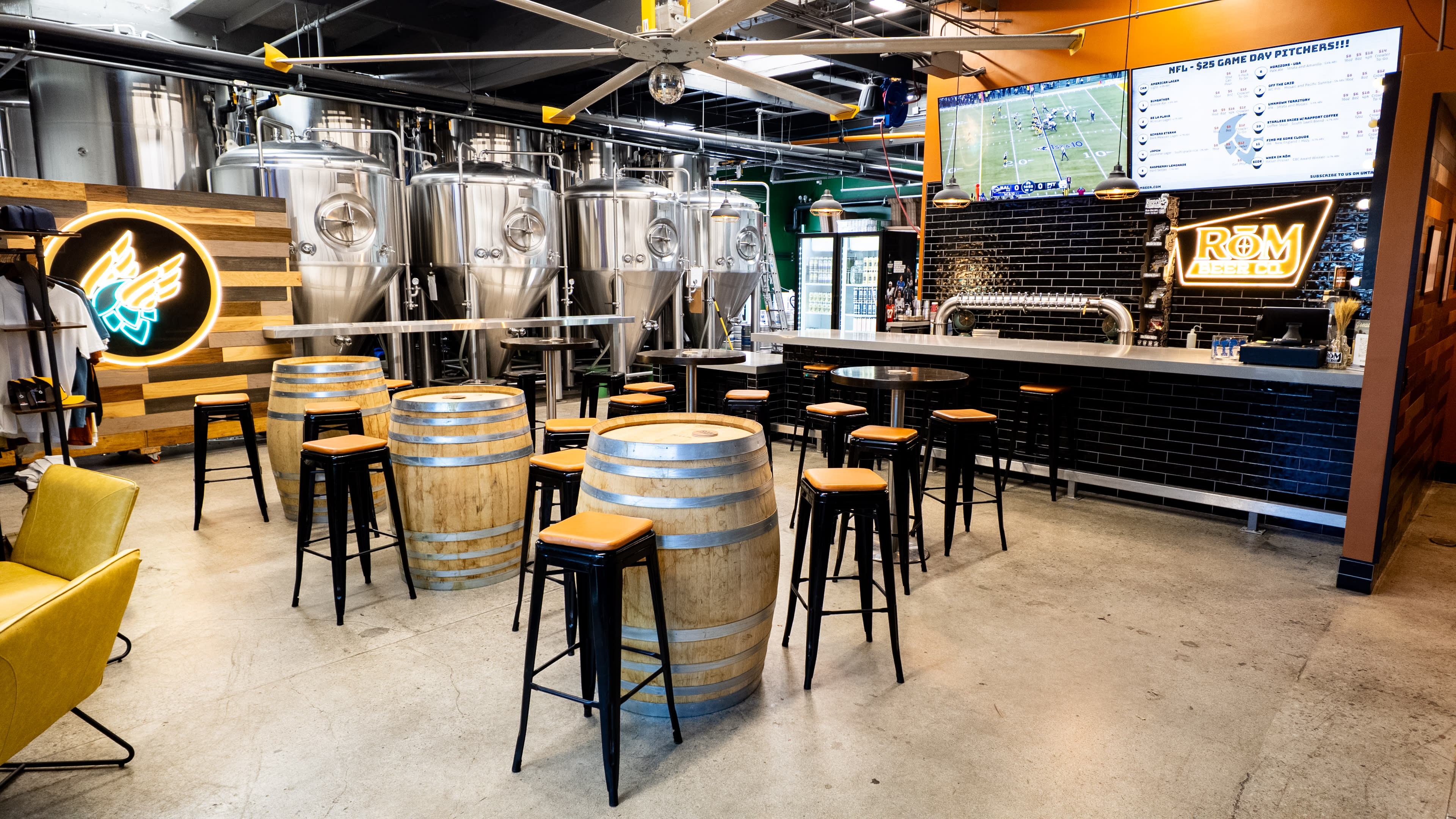 The image shows a modern bar area with wooden barrel tables, high black stools, and large brewing tanks in the background, complemented by digital screens displaying information.