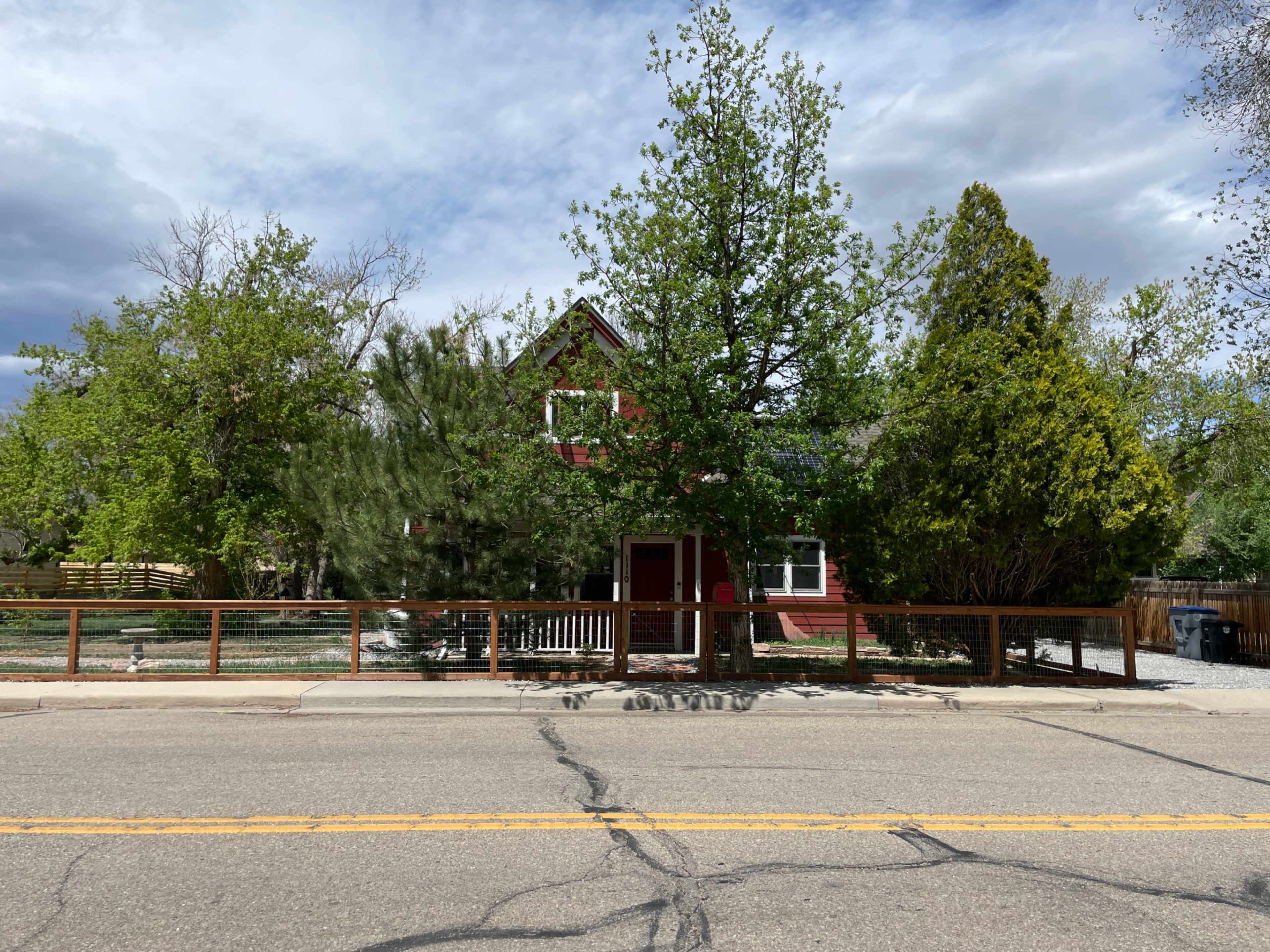 A red house with a porch is surrounded by trees and a wooden fence along a cracked road.
