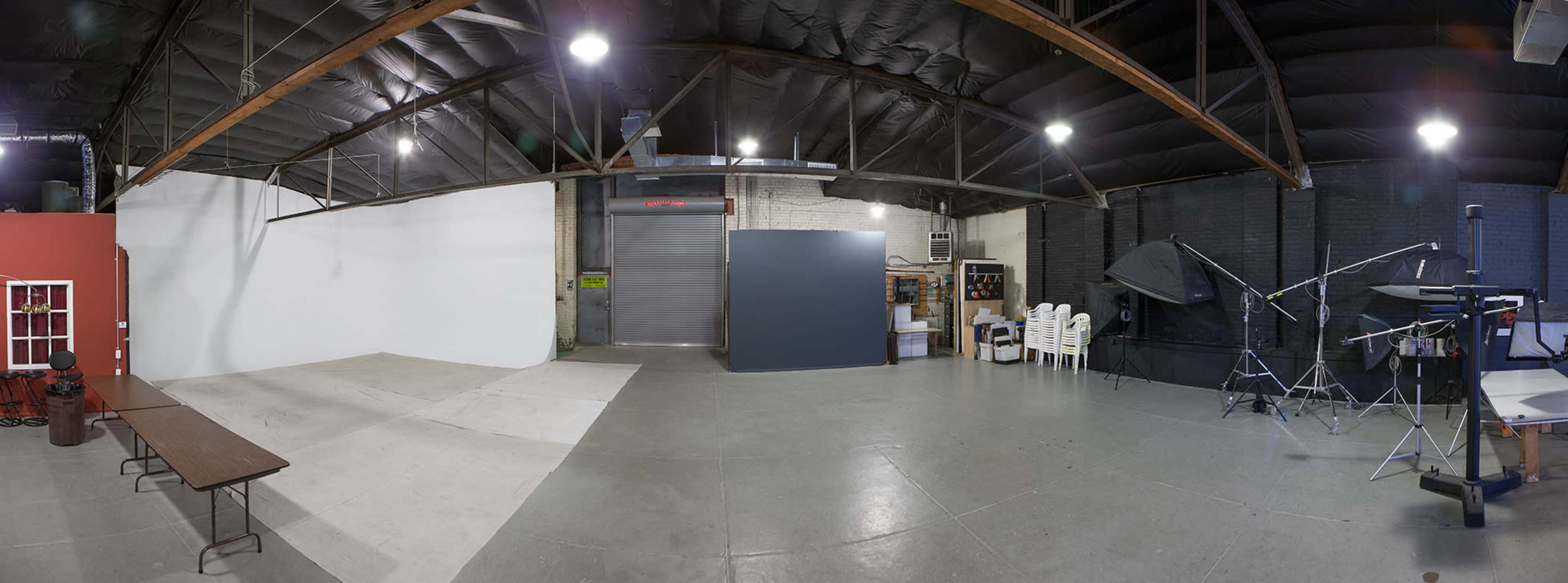 A spacious photography studio with a large white backdrop, a gray wall, several lighting setups, and chairs arranged against a dark wall.