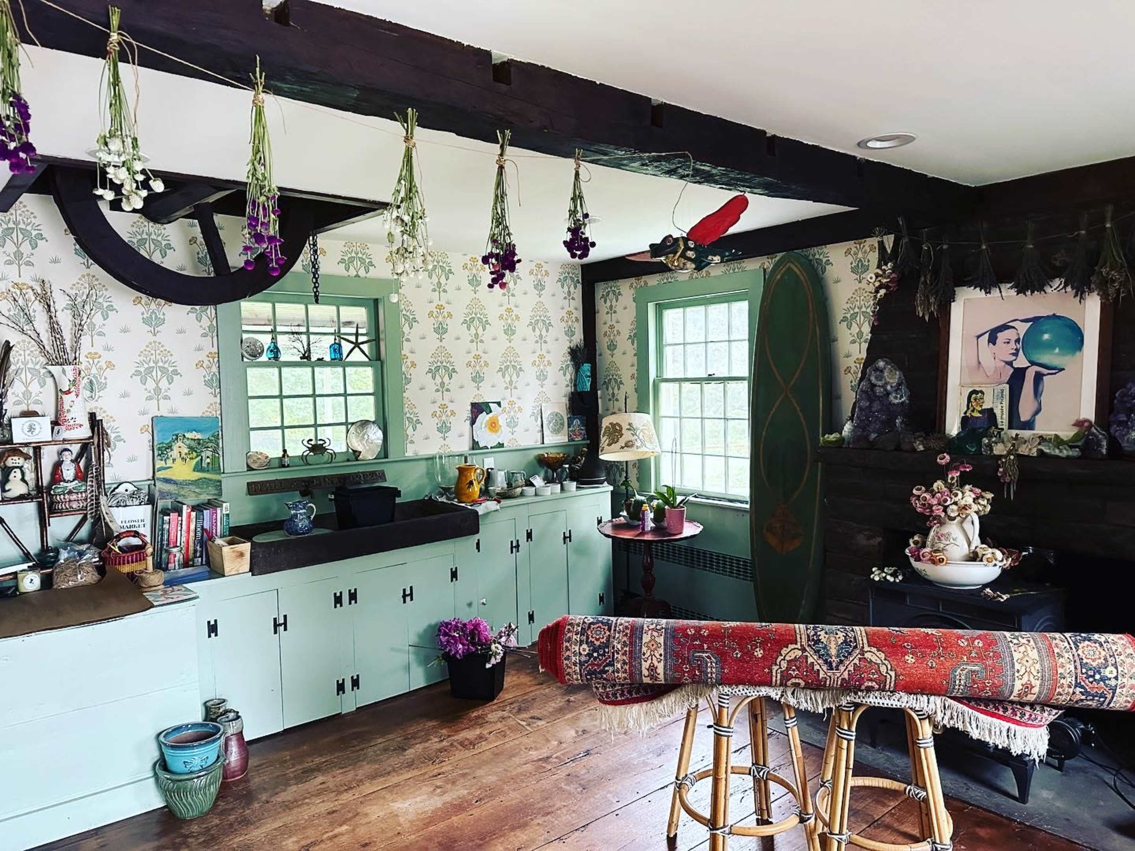 A cozy, eclectic room with green-trimmed windows, hanging dried flowers, and a mix of decorative items on shelves and furniture.