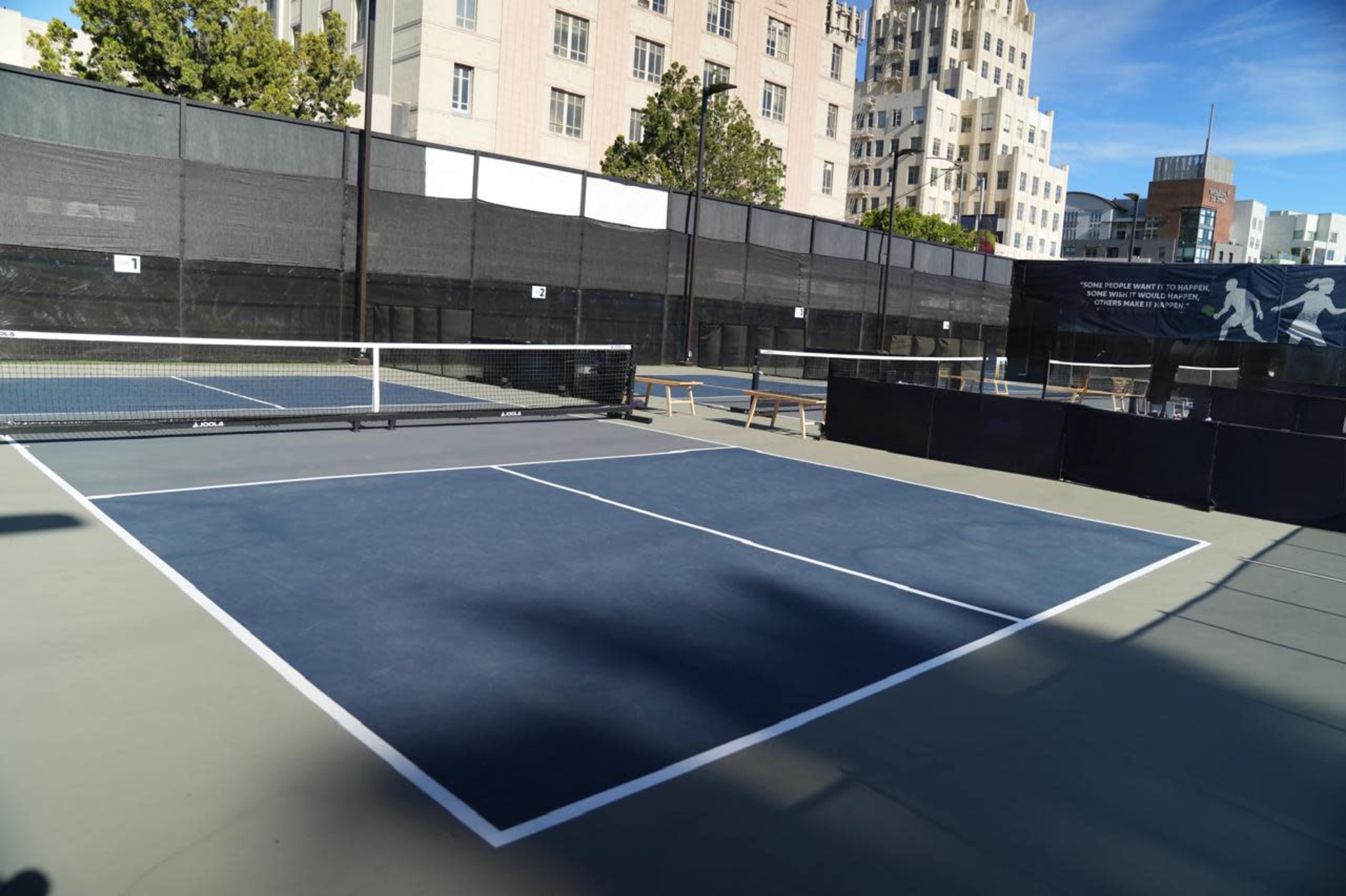 The image shows a tennis court with blue surfaces, surrounded by black fencing and buildings in the background.
