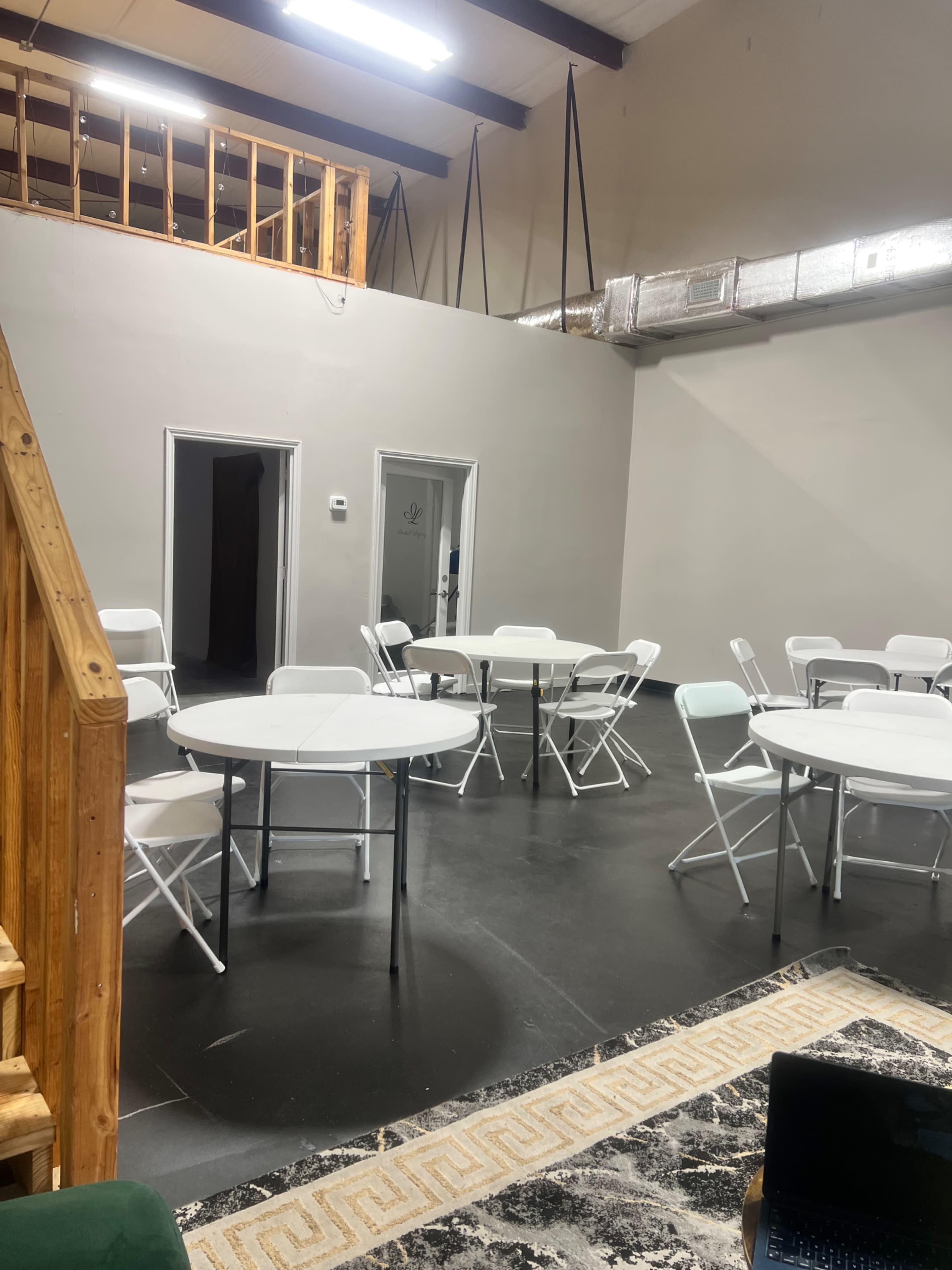 The image shows an interior space with several round tables and chairs arranged, wooden stairs leading to a loft, and an industrial-style ceiling with ductwork.