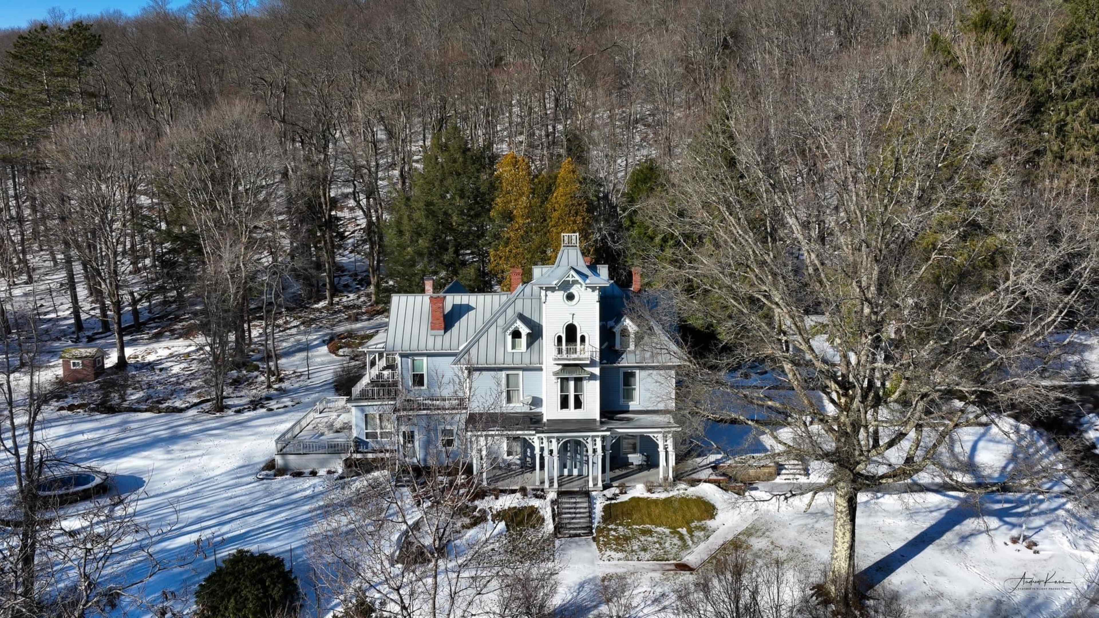 A large, historic house with a steep roof and multiple gables sits in a snowy landscape, surrounded by bare trees.